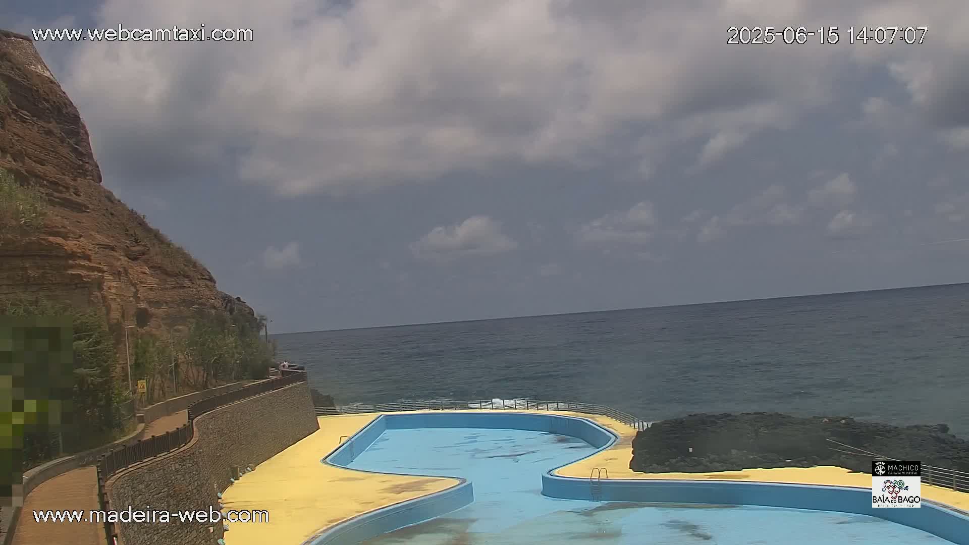 A partly cloudy day shows an outdoor pool adjacent to a rocky cliff overlooking a calm ocean.