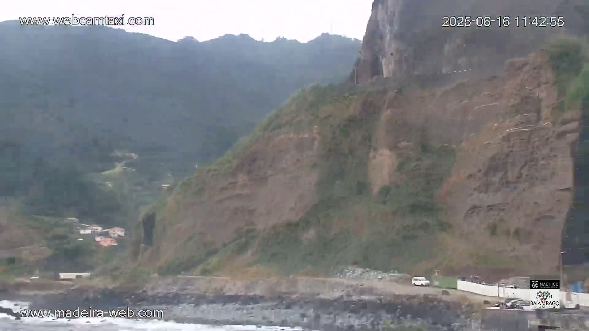 A coastal road winds along a rocky cliff face overlooking a calm ocean, under an overcast sky.