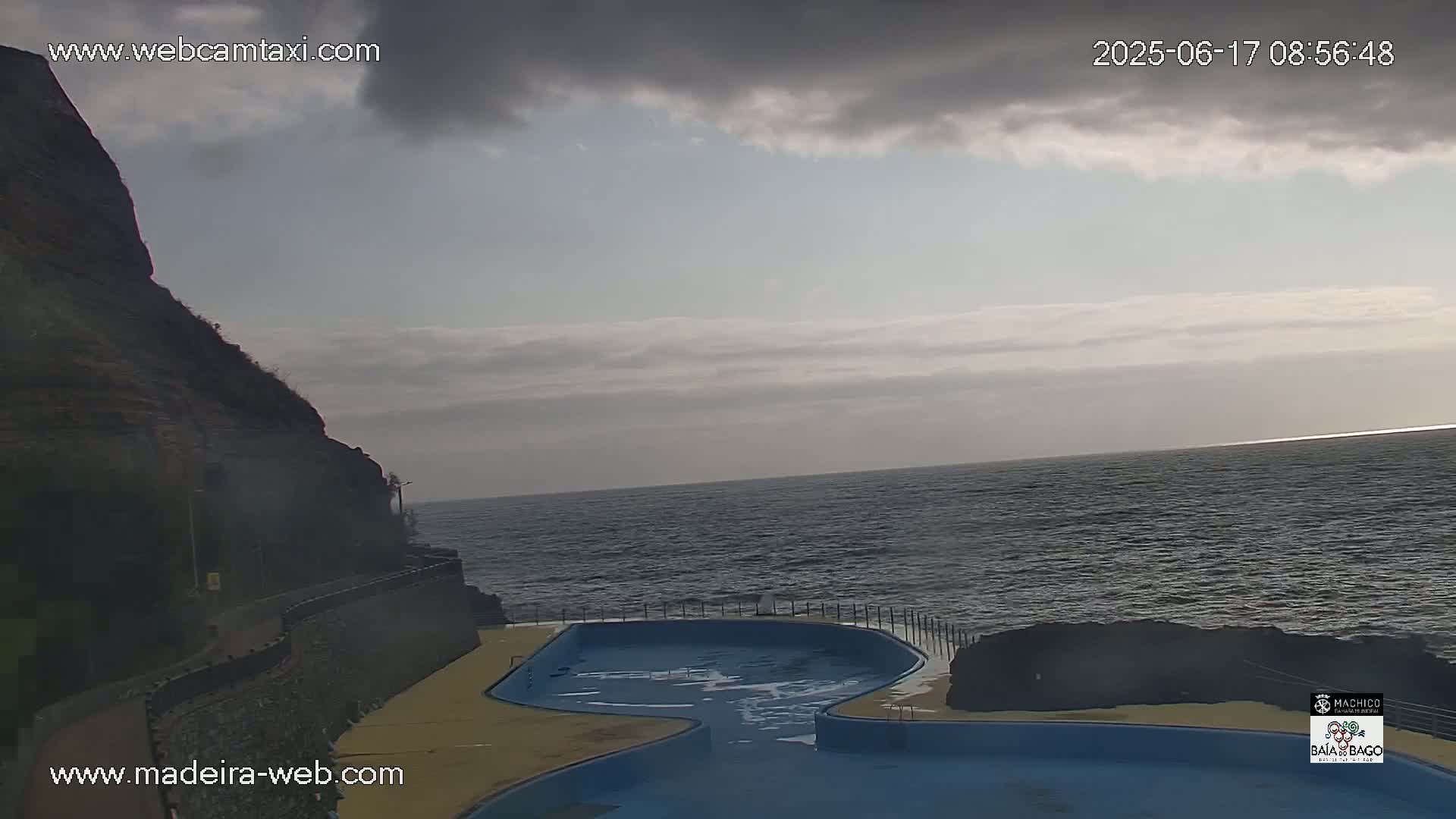 An outdoor scene shows a partially cloudy sky over a dark-blue ocean, with a rocky cliff to the left and a blue pool next to the water.