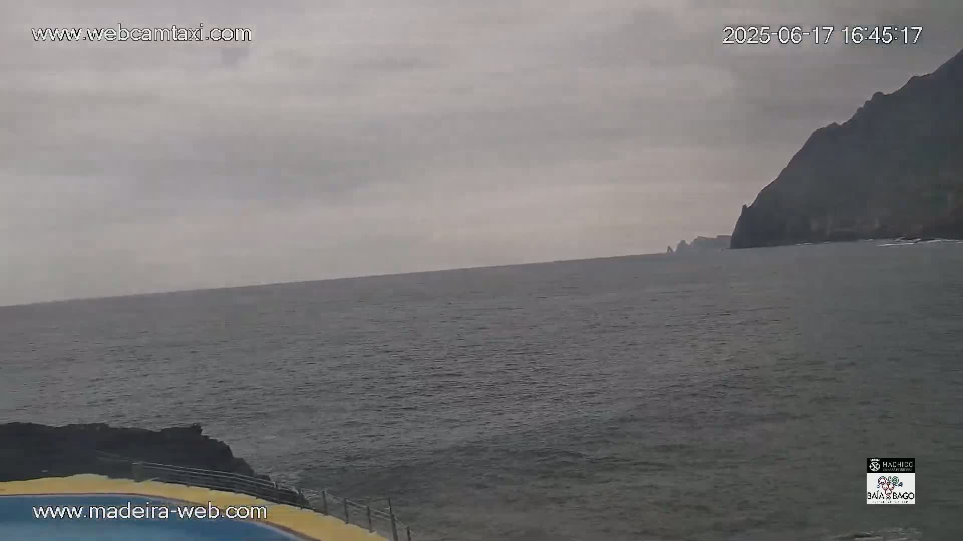 A dark, overcast sky hangs over a calm, gray ocean meeting a dark cliff face in the distance.