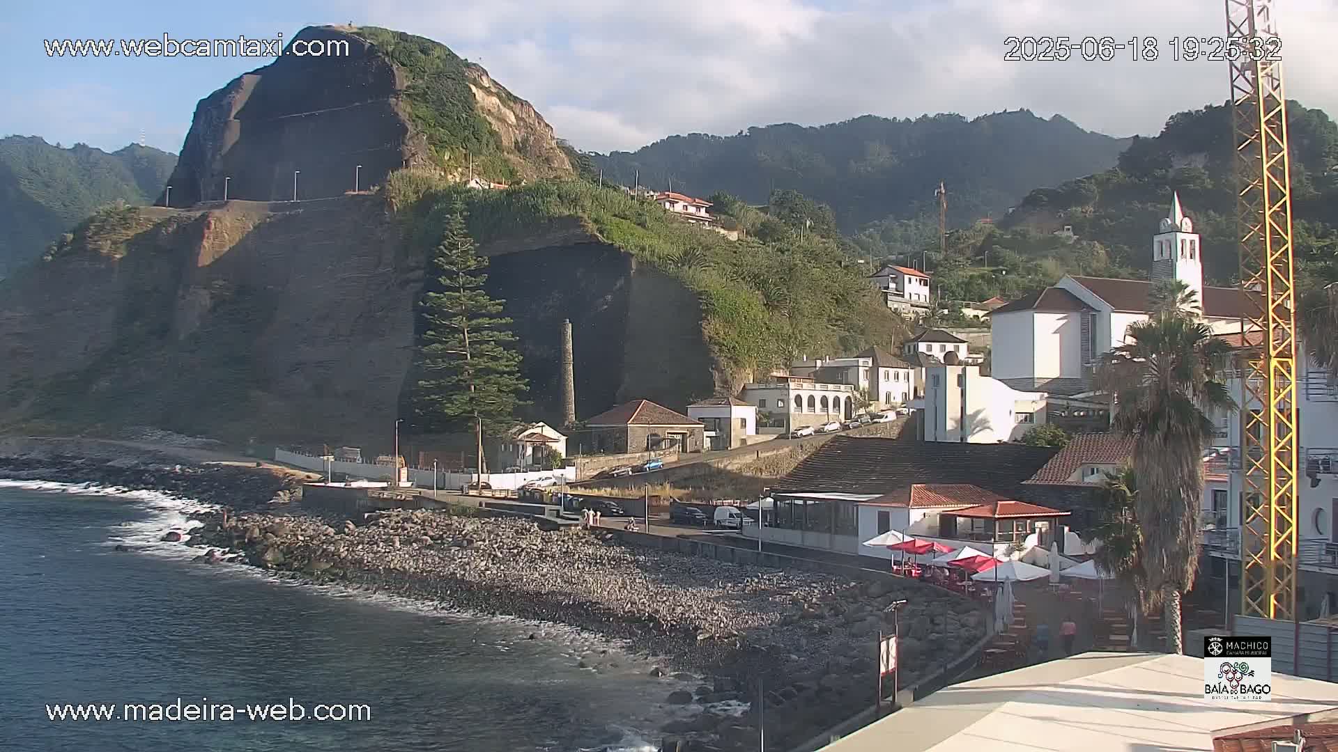 A coastal town nestled against a steep, dark cliff overlooking a calm ocean under a mostly clear sky.