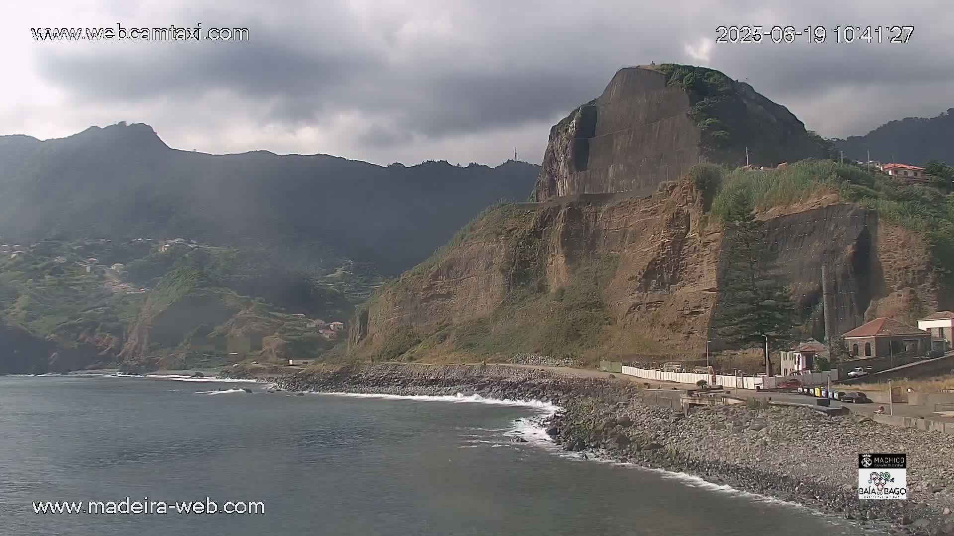 A rocky coast with a steep cliff face and a calm ocean under a mostly cloudy sky.