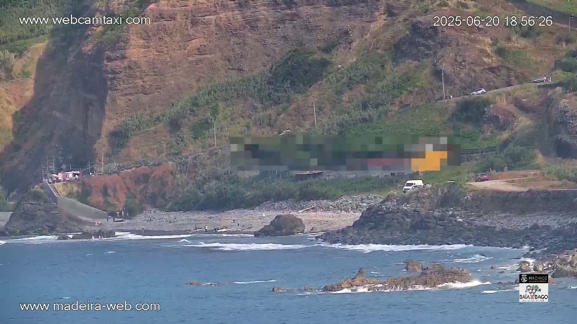 A rocky, dark-colored coastline with a small beach features a few people, some vehicles on a road above, and a slightly wavy ocean under a sunny sky.