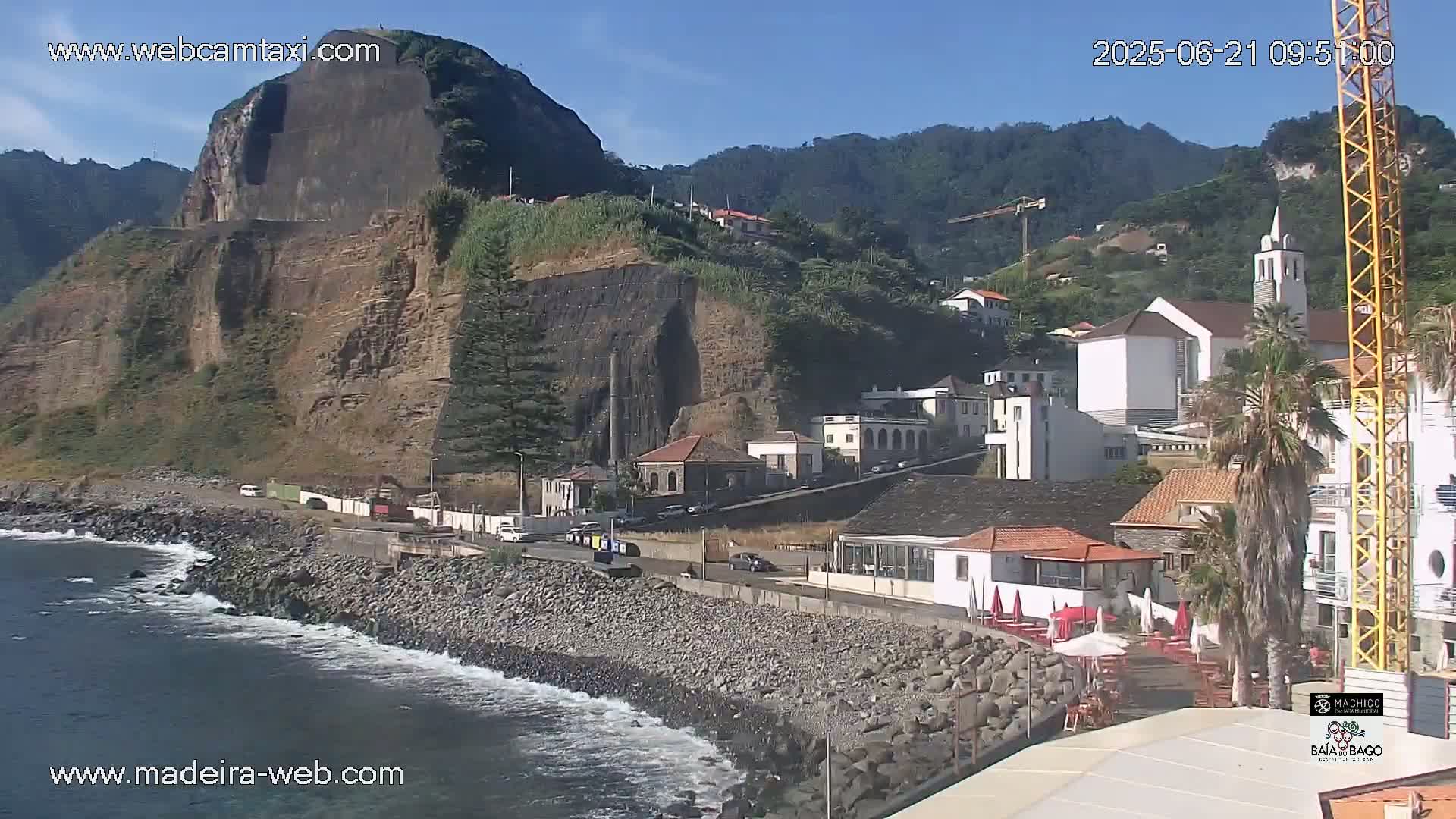 A rocky coastline with a village nestled against a steep, green hillside under a clear blue sky.