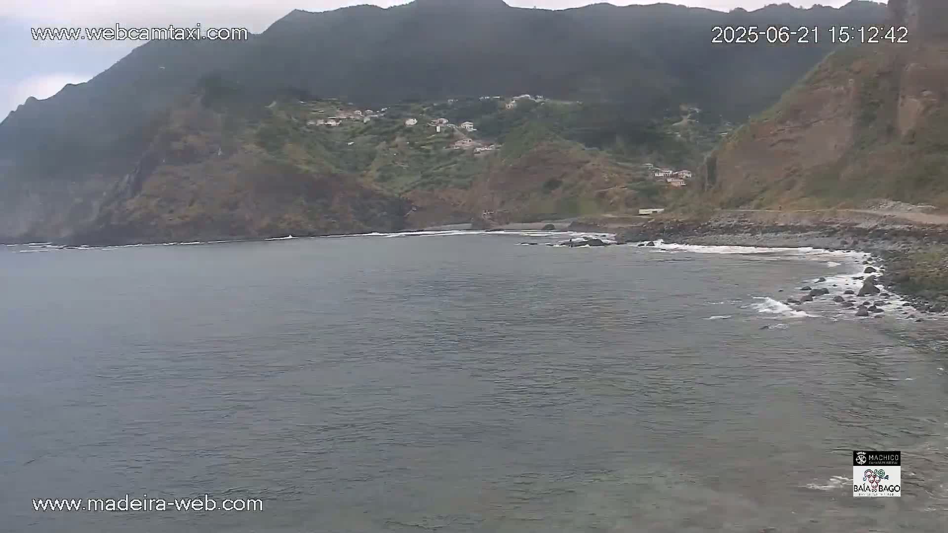 A rocky, dark-colored coastline with a small beach curves into a calm, grey ocean under an overcast sky.