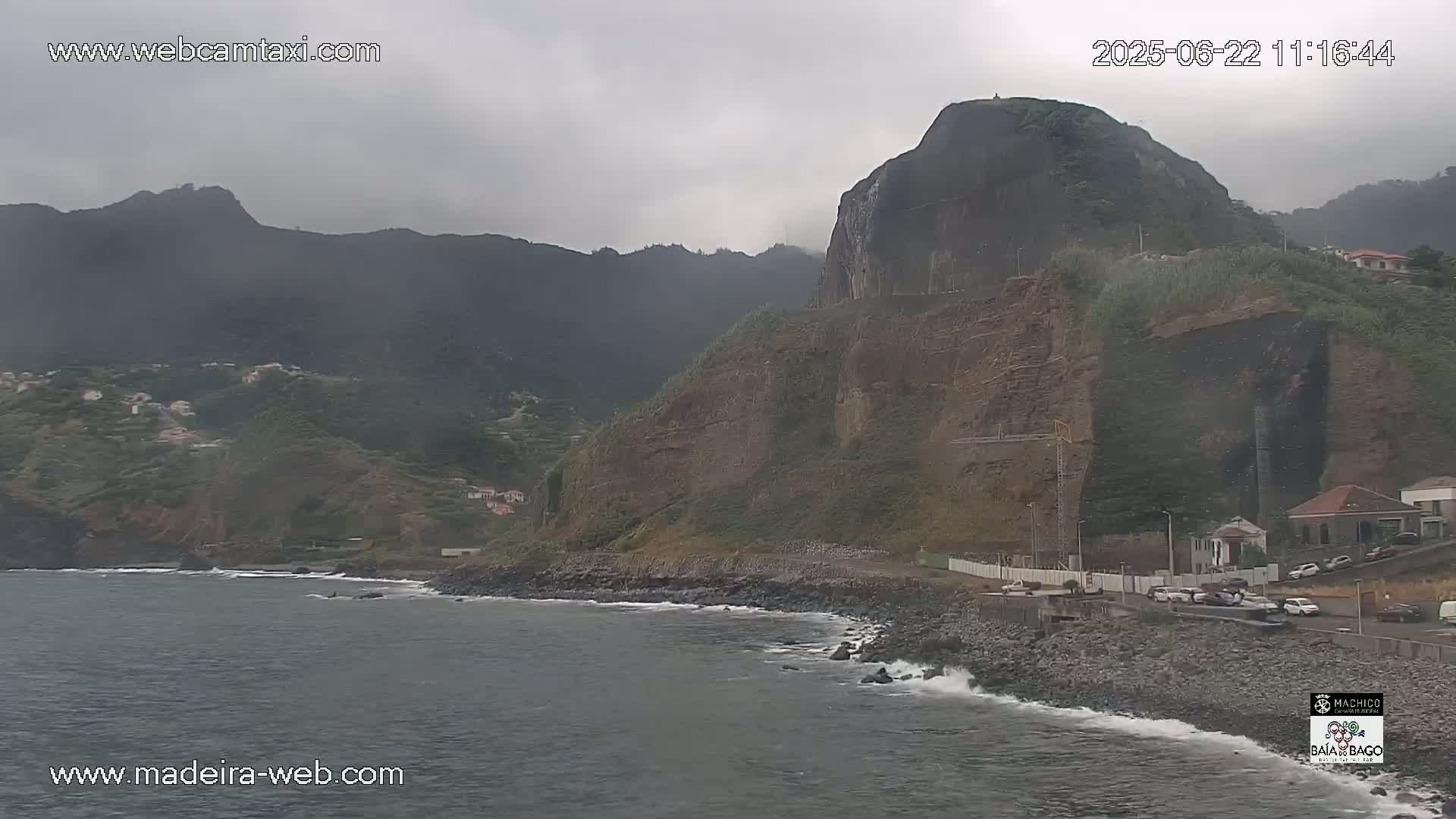 A rocky coastline with dark gray mountains in the background under an overcast sky shows a small town along the water's edge with some small waves crashing on the shore.