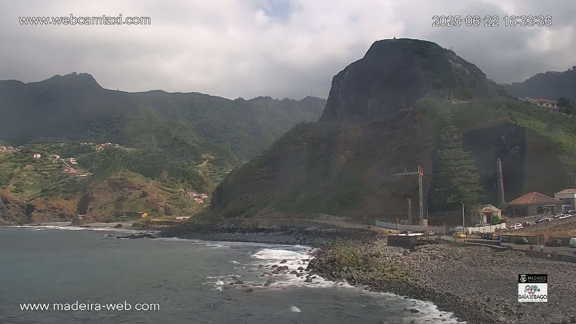 A rocky coastline with a dark, steep mountain in the background under overcast skies, shows light waves breaking on a pebble shore.