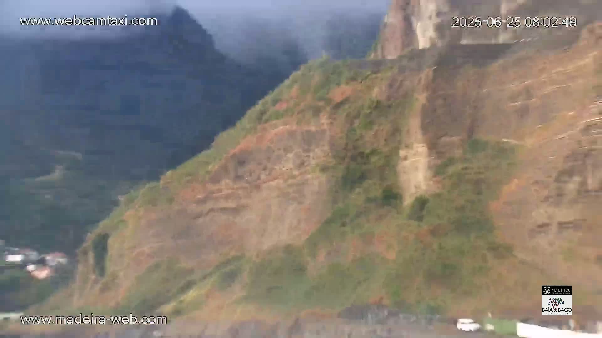 A hazy view shows a steep, rocky cliff face partially covered in vegetation, overlooking a small village below.