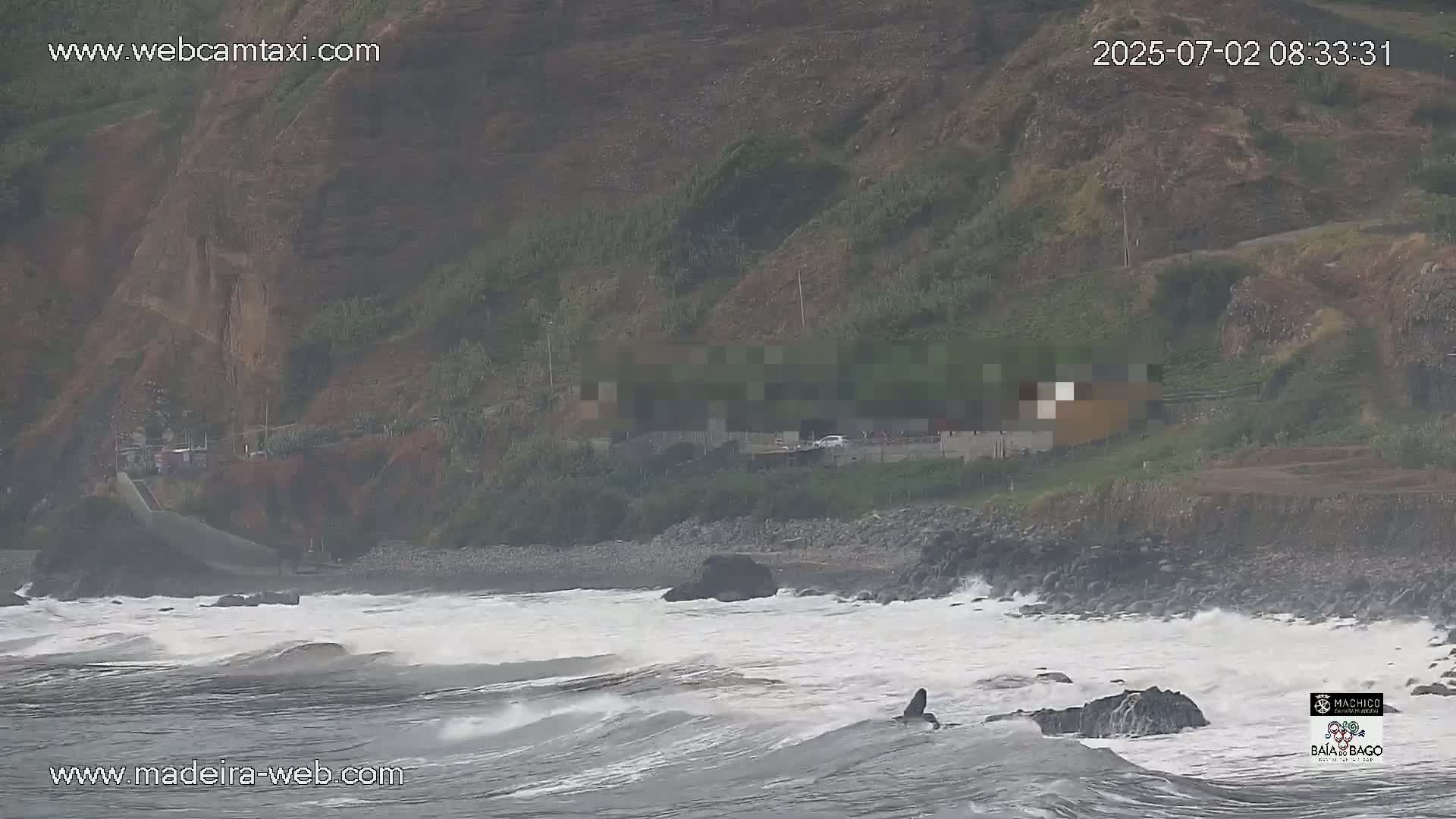 Rough waves crash against a rocky shoreline below a hillside with sparse vegetation in overcast conditions.