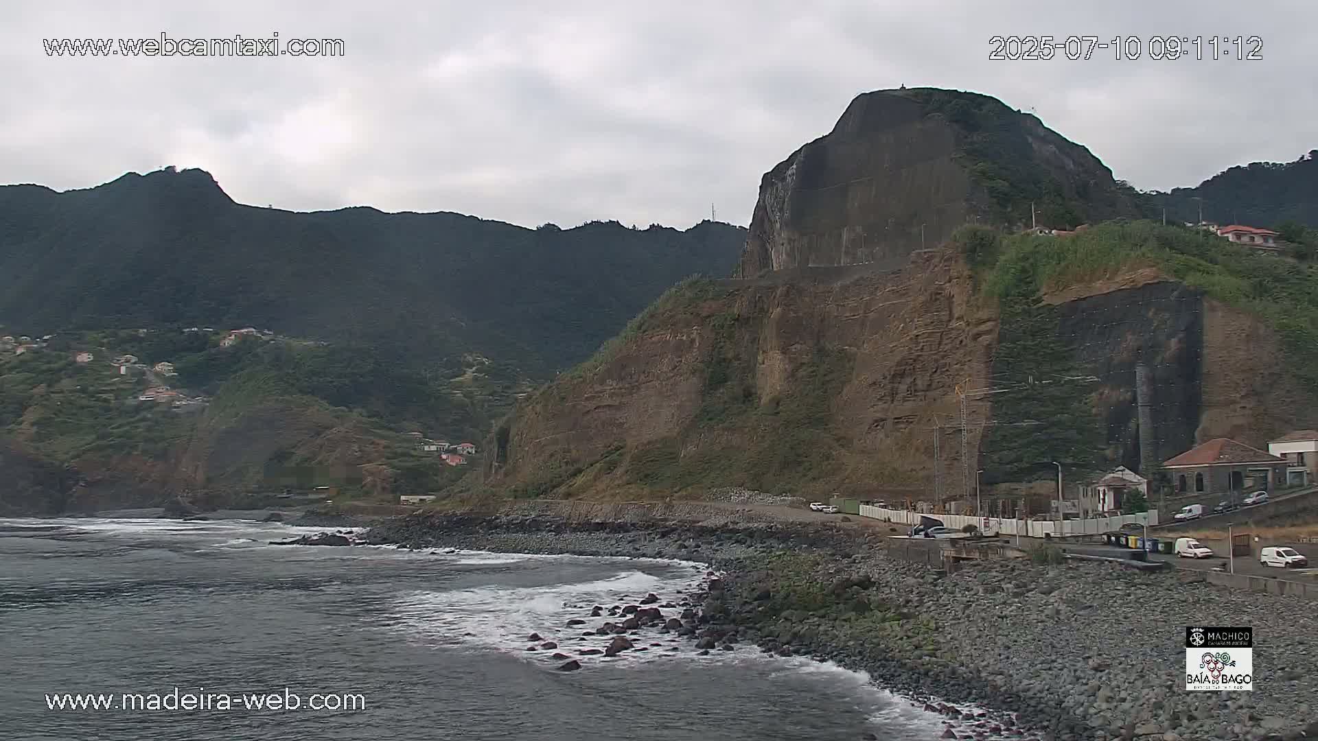 A rocky coastline with dark, grey-brown cliffs and a large, flat-topped rock formation overlooking a dark-grey ocean with small waves under an overcast sky.