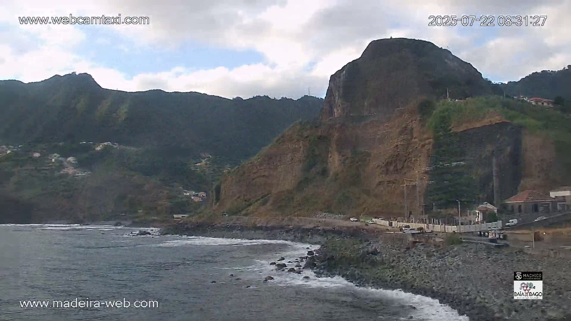 A rocky, mountainous coastline with a partly cloudy sky meets a dark-grey ocean with small waves.
