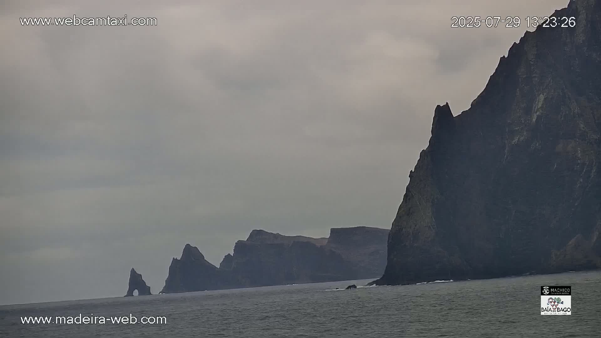 A rocky, mountainous coastline with a partly cloudy sky meets a dark-grey ocean with small waves.
