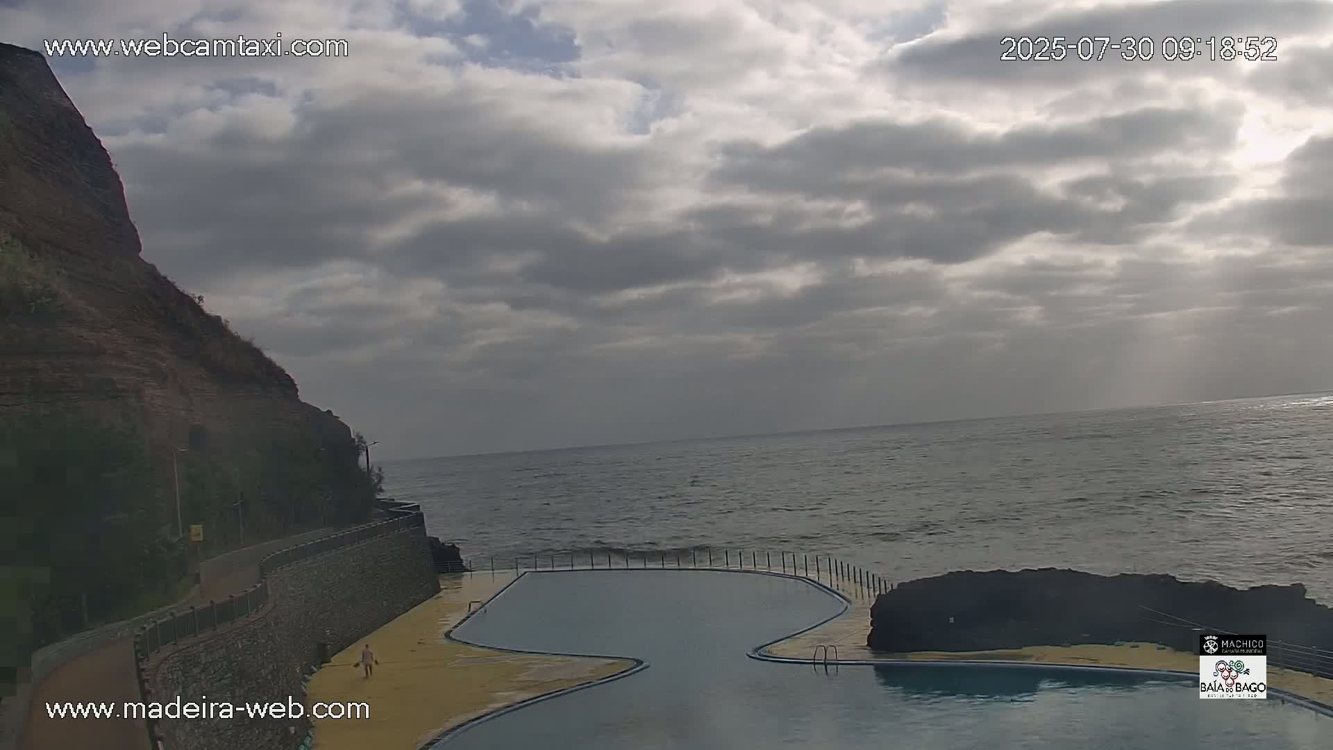 A partially cloudy sky overlooks a coastal scene featuring a curved oceanfront pool and a rocky cliff.