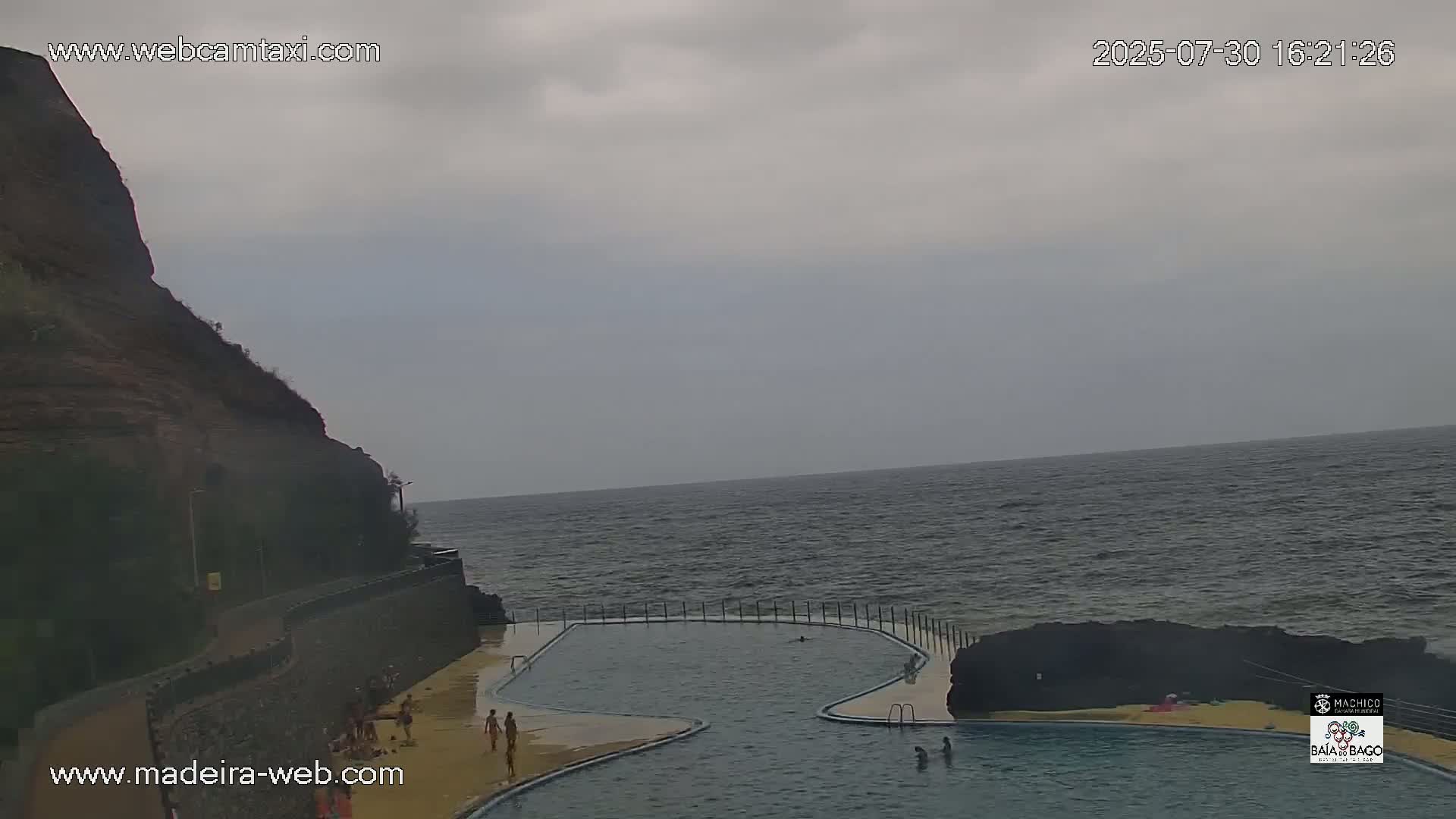 A partially cloudy sky overlooks a coastal scene featuring a curved oceanfront pool and a rocky cliff.