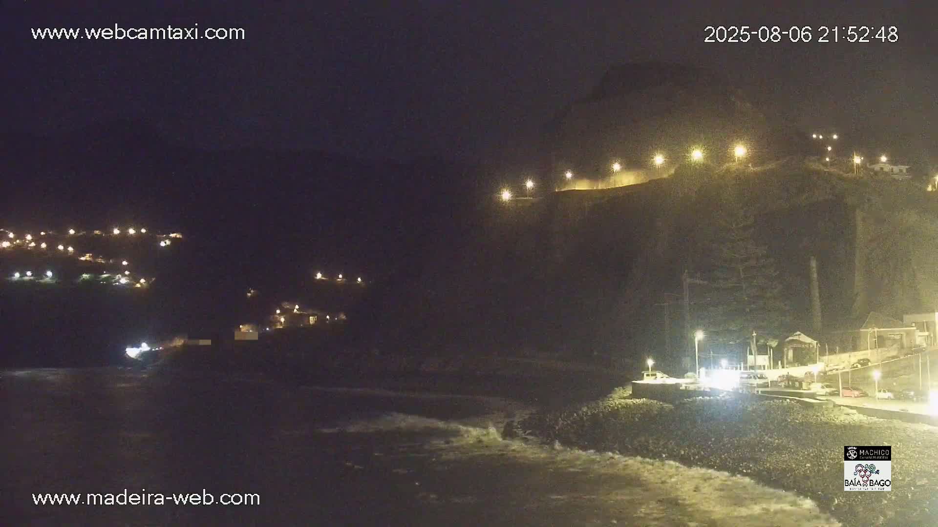 A nighttime coastal scene shows a dark cliff face with lights illuminating a roadway along its top, a small town nestled into the hillside below, and waves lapping at a shoreline.