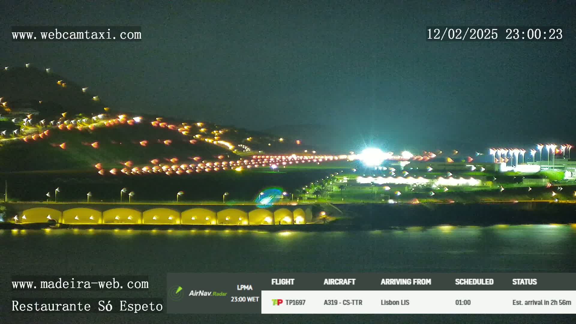 A nighttime long shot of a hillside with numerous lights, a waterfront structure with lights, and an airport runway with lights, all under a dark sky.