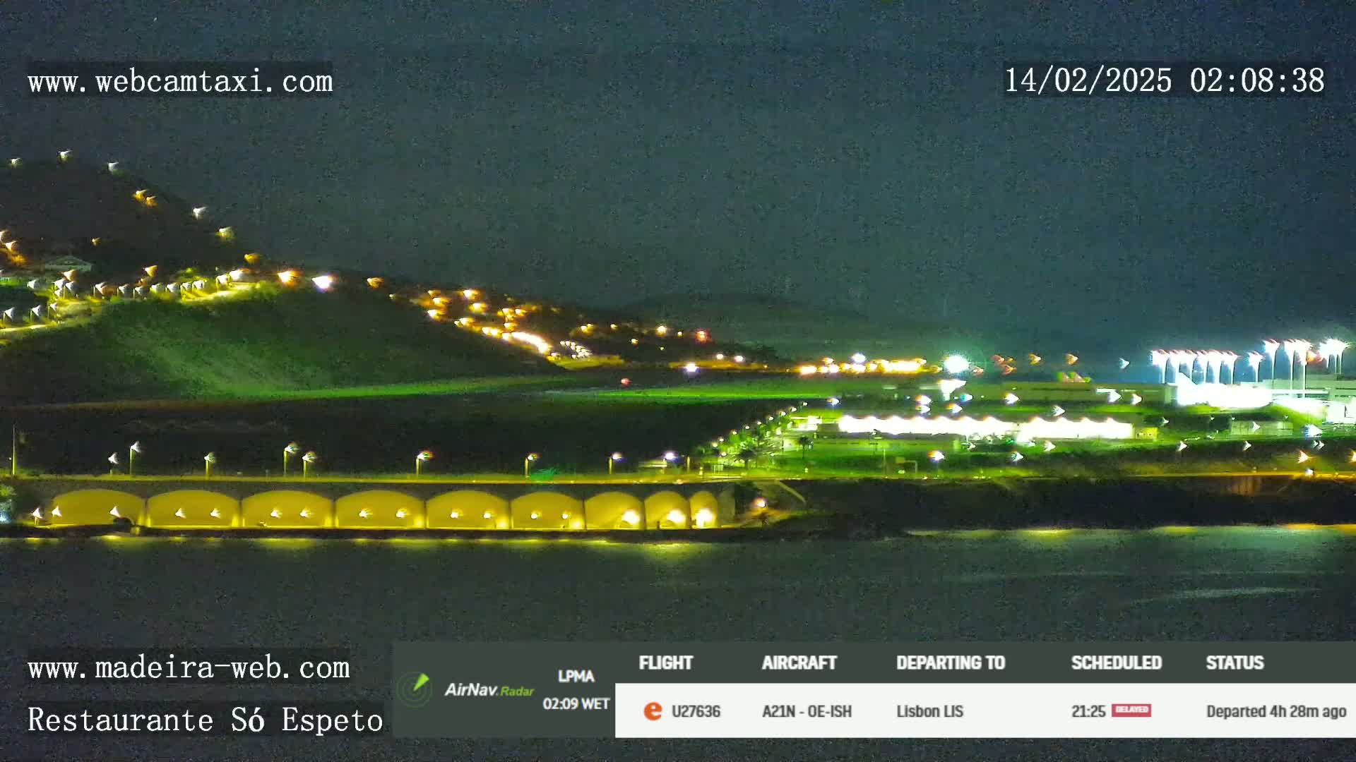 A nighttime coastal scene shows a hilltop with illuminated buildings, a waterfront area with lights, and a dark body of water under a clear night sky.