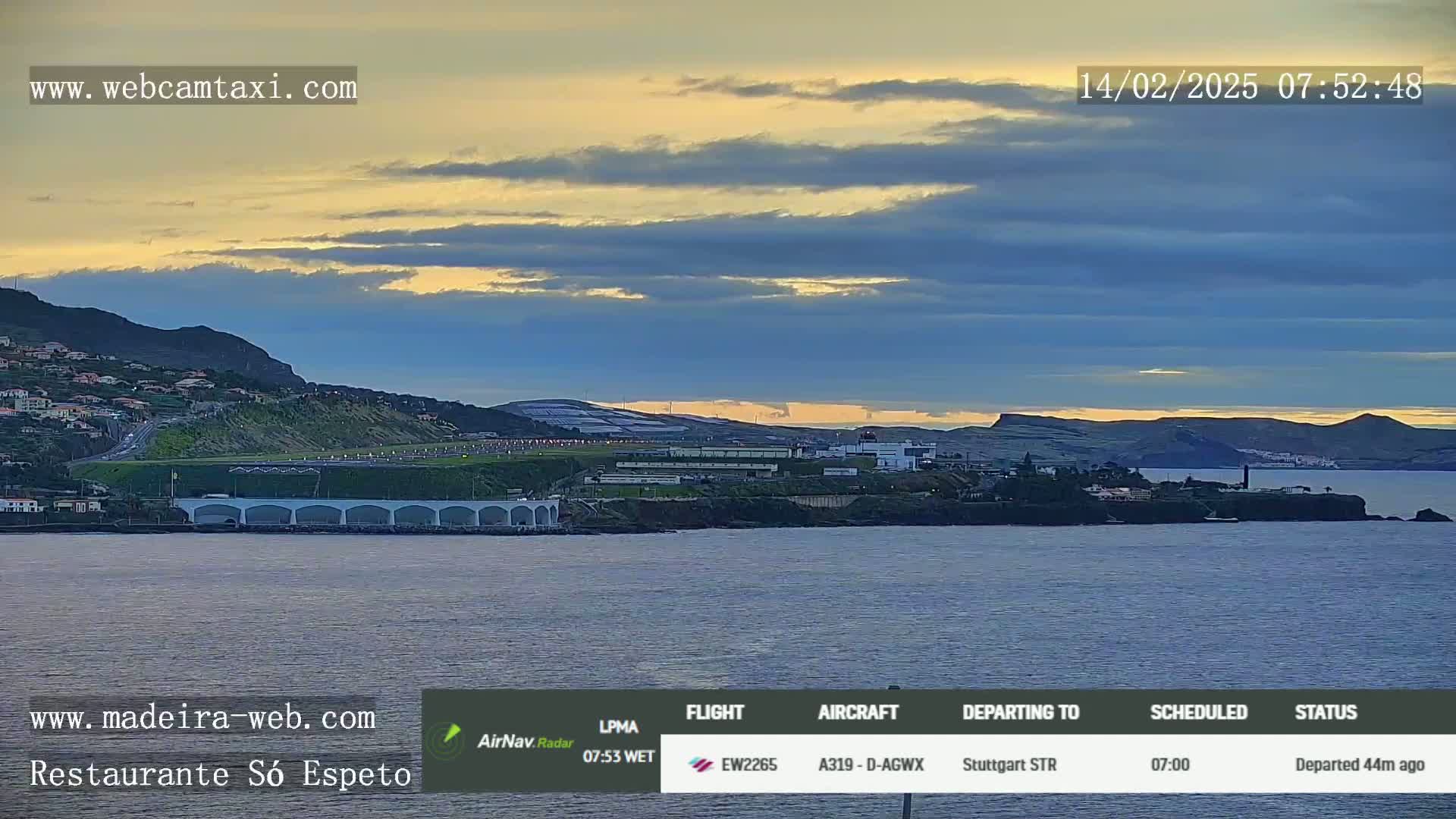 A coastal scene at sunrise shows a body of water in front of a hilly landscape with buildings, an airport, and a bridge.