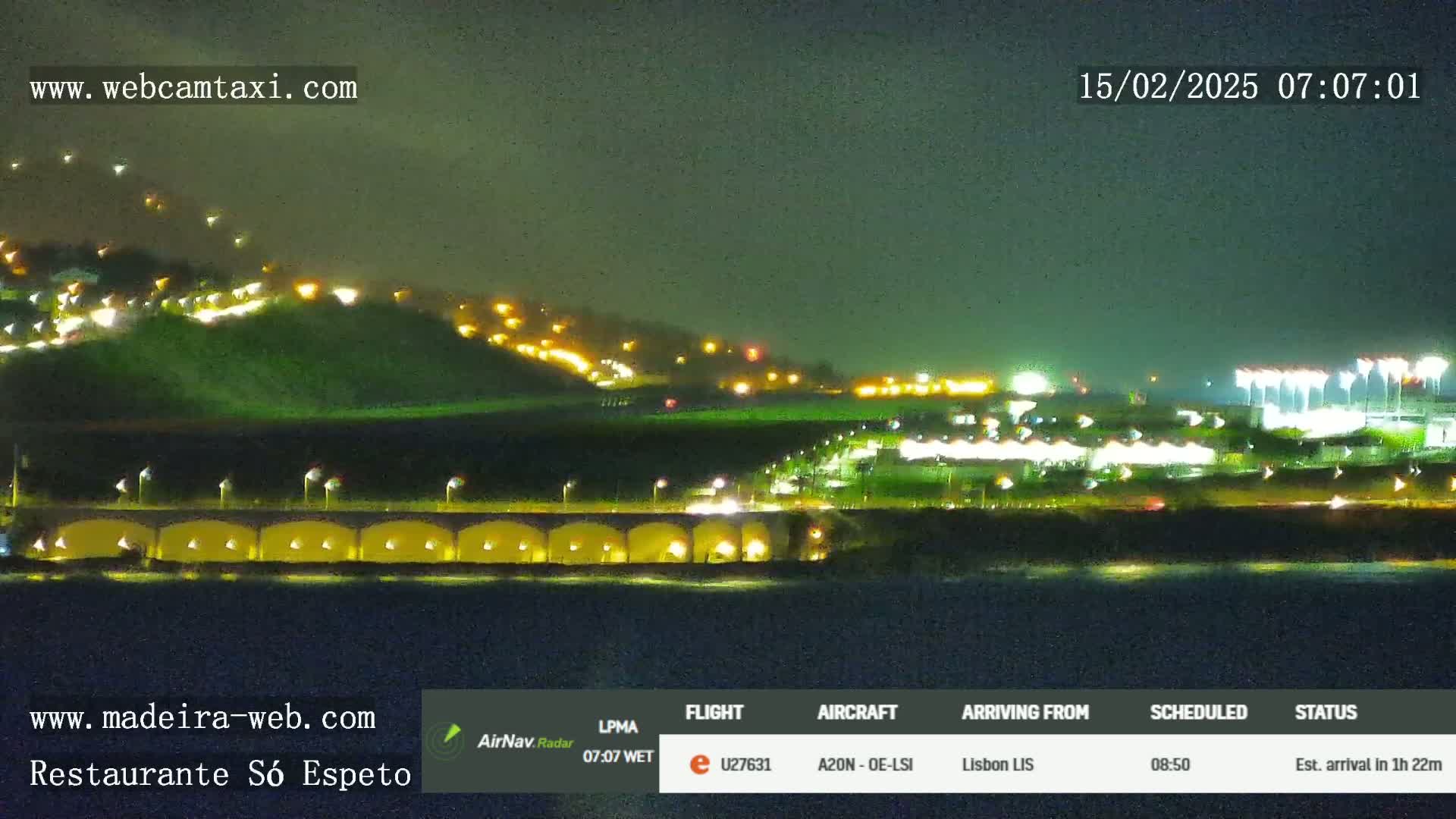 A nighttime, dimly lit coastal scene shows a hill with scattered lights, a long structure with arched openings and lights, and a body of dark water in the foreground.