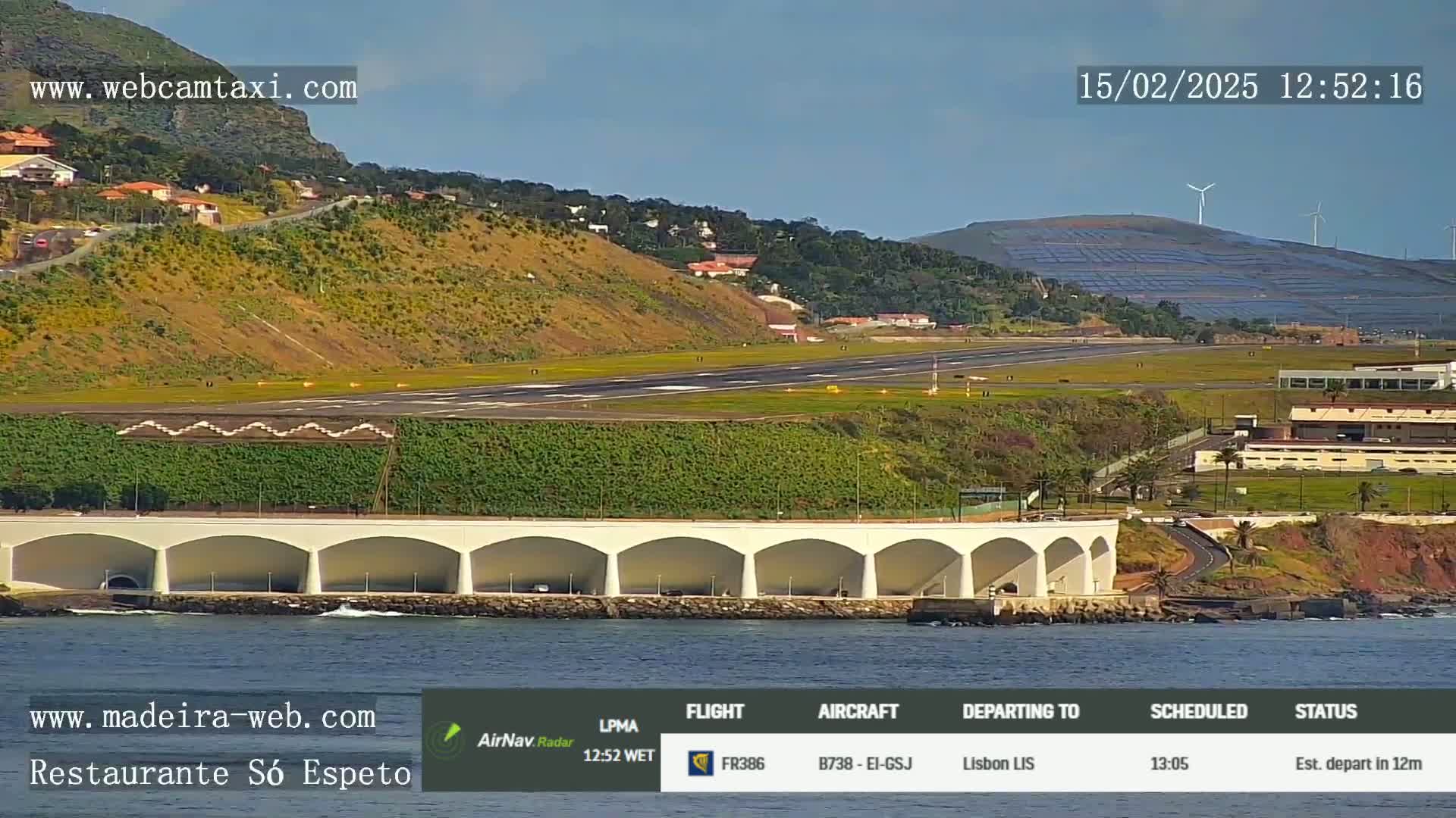 An airport runway is visible, situated near a coastline with calm ocean waters, under a clear sunny sky; a hillside with wind turbines is in the background.