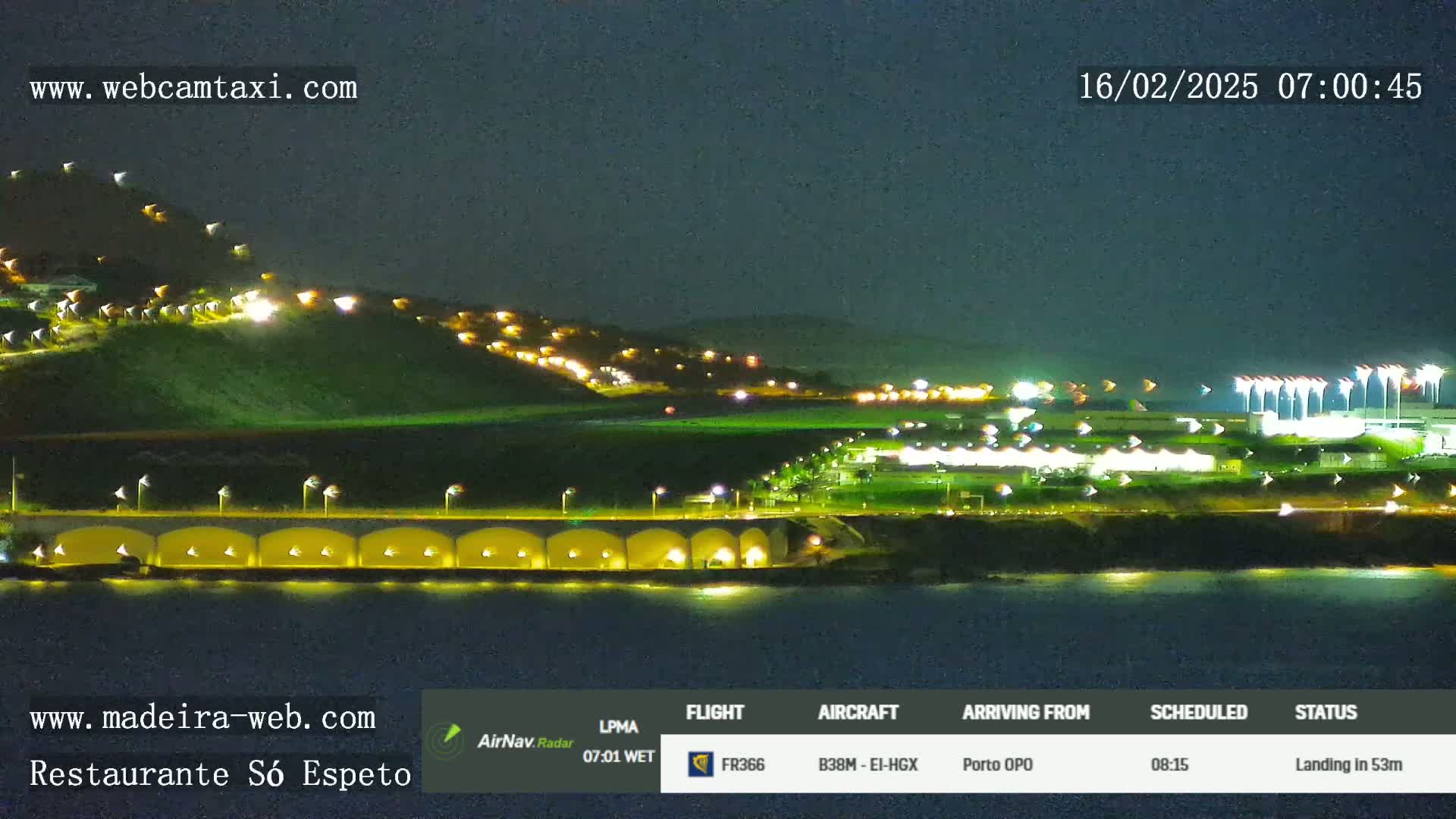 A nighttime view of a coastal town with illuminated buildings on a hill overlooking a dark body of water, under a clear night sky.