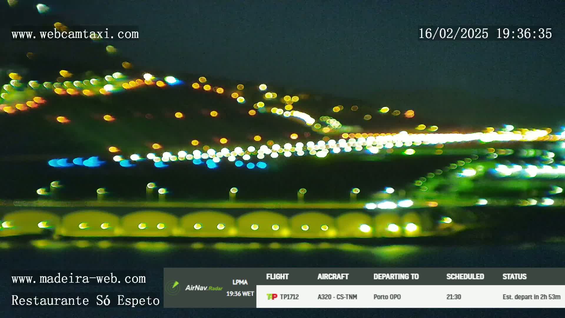 A nighttime view of a hilltop with numerous blurry lights, a body of water with lights reflecting on it in the foreground.