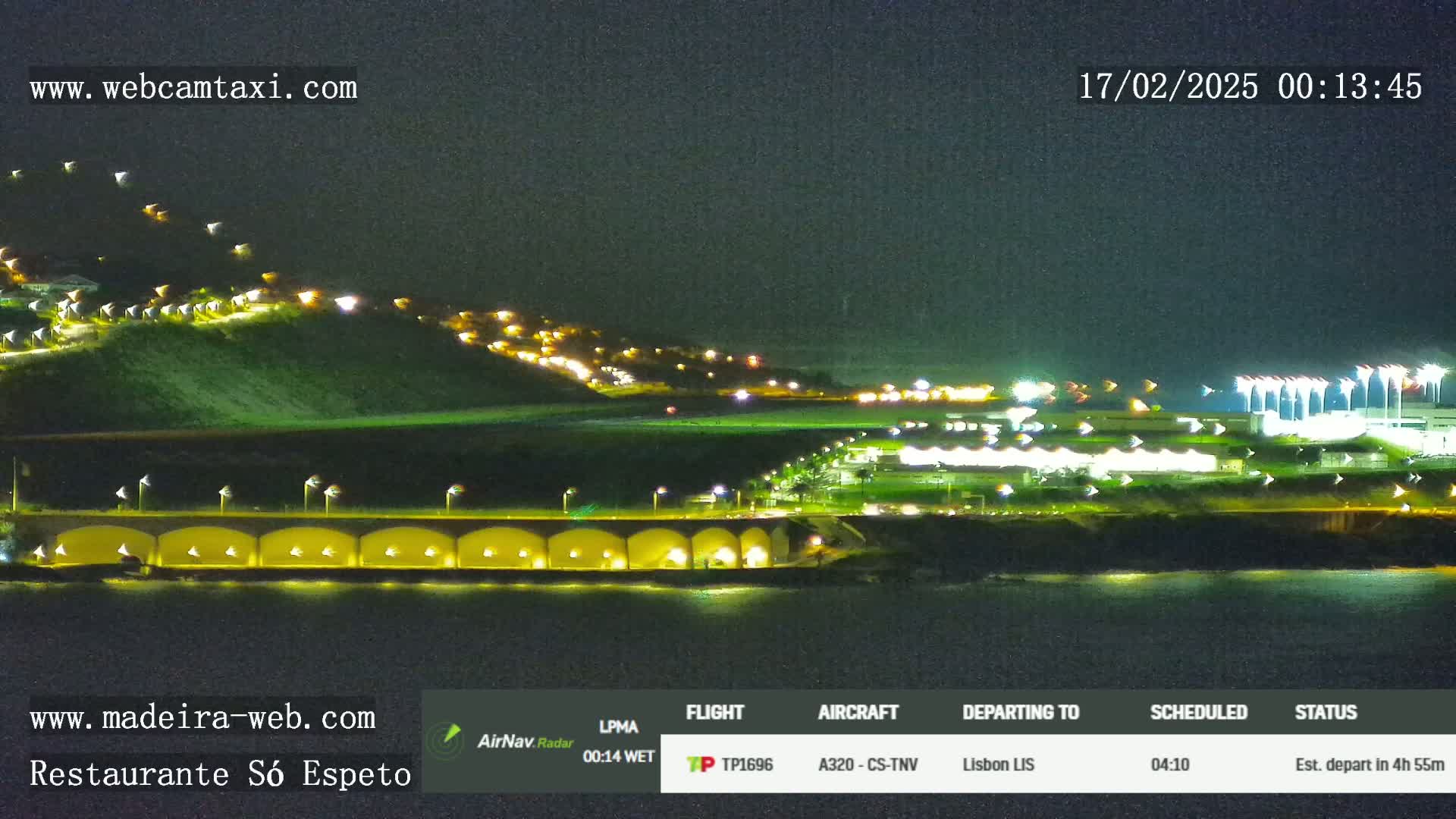 A nighttime coastal scene shows a hilltop with scattered lights, a long, low structure with lights along a waterfront, and an airport runway with lights in the distance.