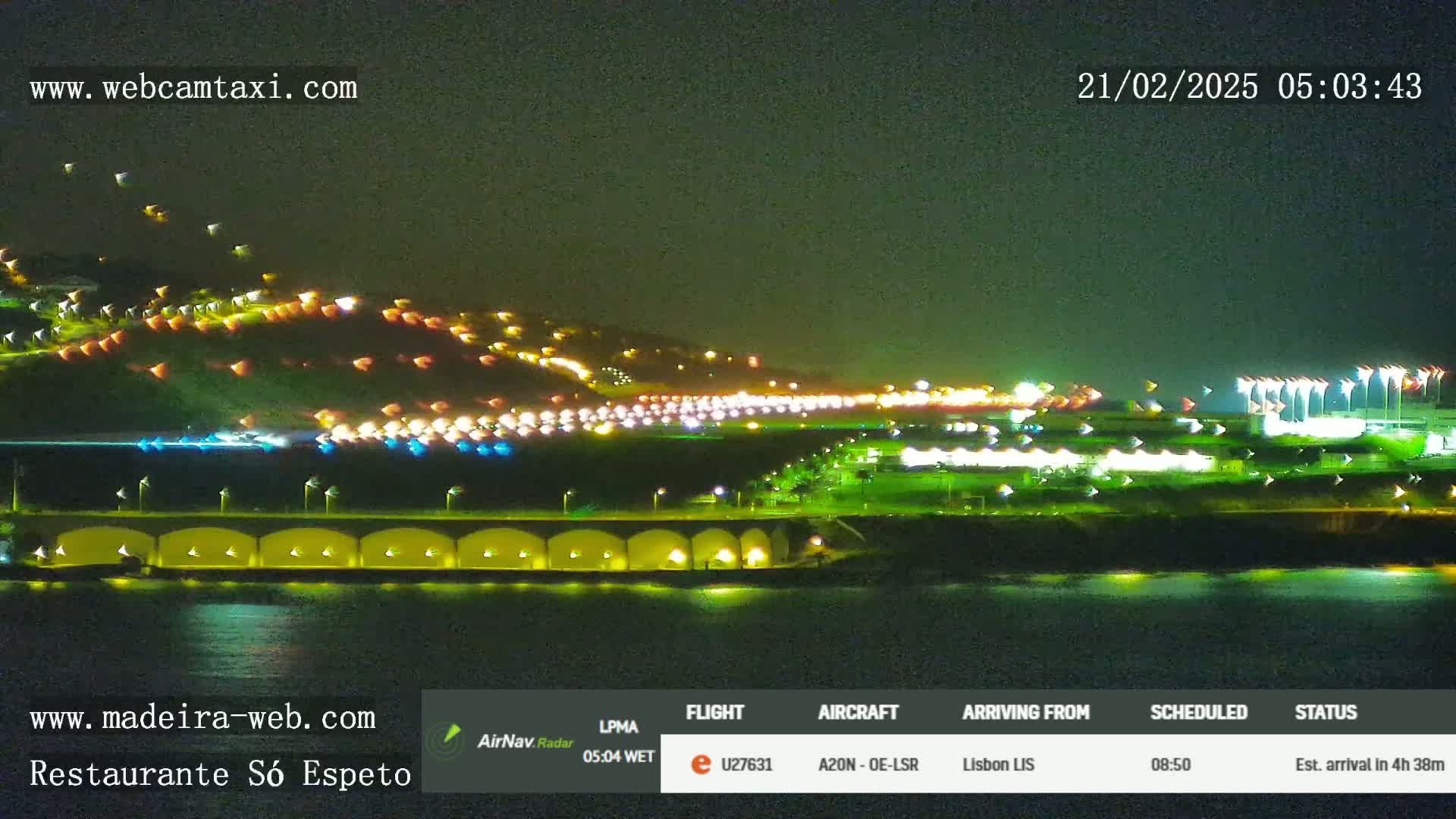 A nighttime coastal scene shows a hillside dotted with lights, a long, brightly lit roadway, and a body of dark water.