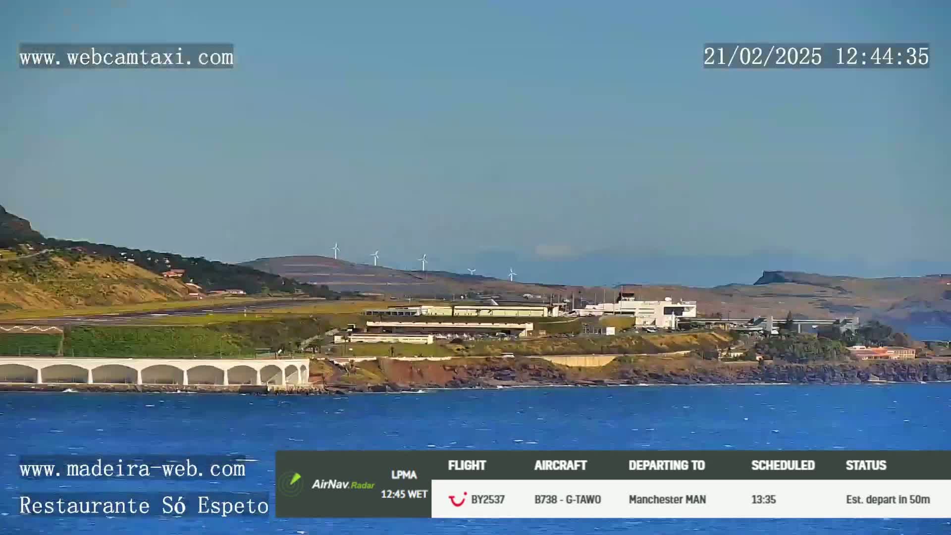 A coastal airport with wind turbines on a hill in the background is viewed from across calm ocean water on a clear day.
