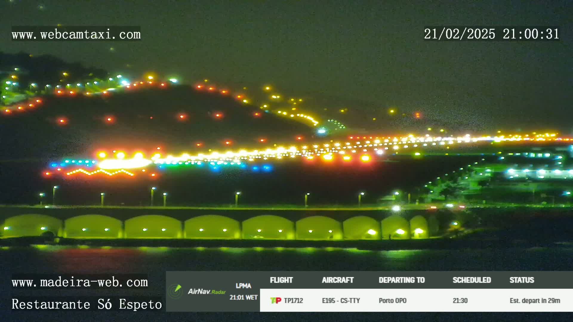 A brightly lit airport runway at night is visible from across a body of water, with hills illuminated in the background.