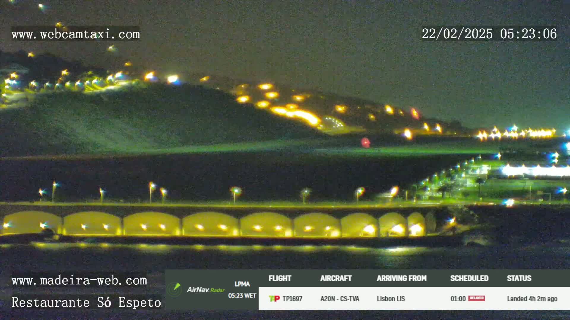 A nighttime coastal scene shows a hill illuminated with many lights, a bridge with lights under it, and dark water.