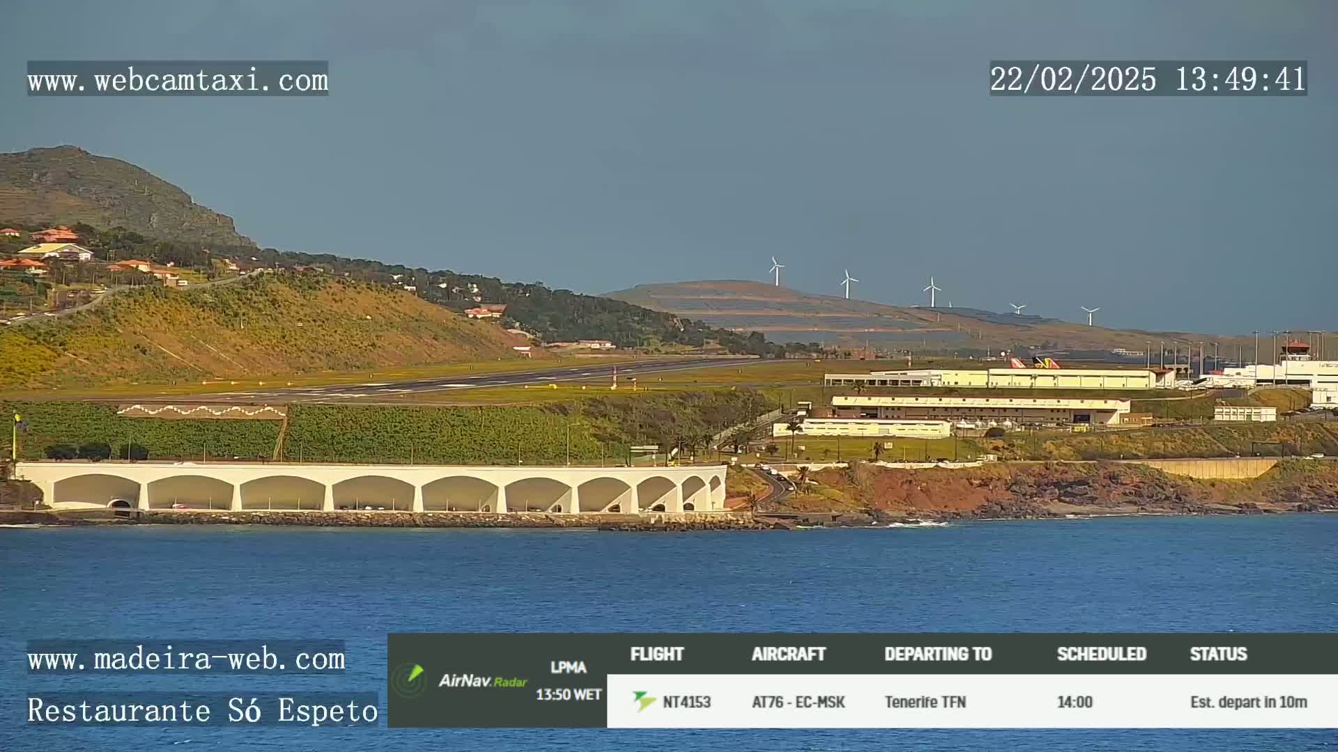 An airport runway, terminal buildings, and wind turbines are visible on a hill overlooking the ocean under a clear sky.