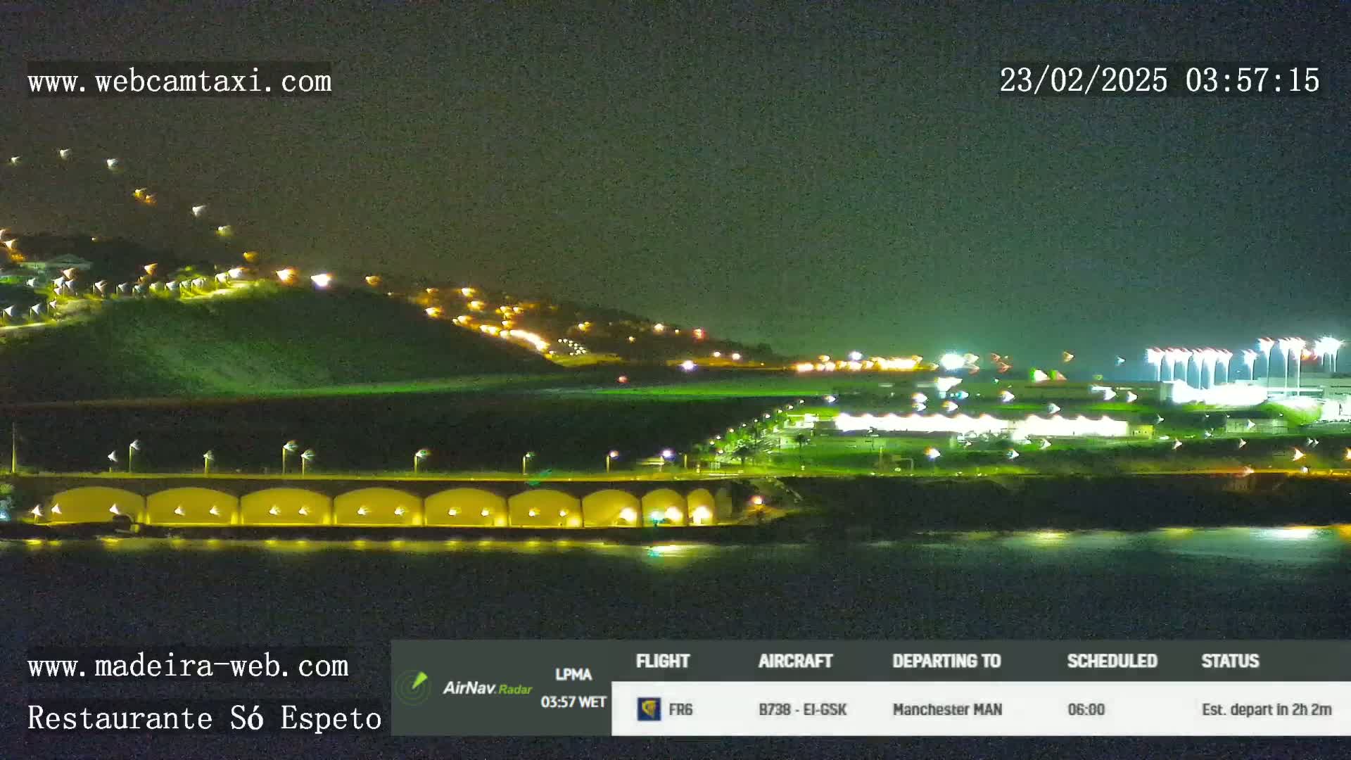 A nighttime coastal scene shows a hill with illuminated buildings, a waterfront structure with arched lights, and a brightly lit airport or stadium in the background.