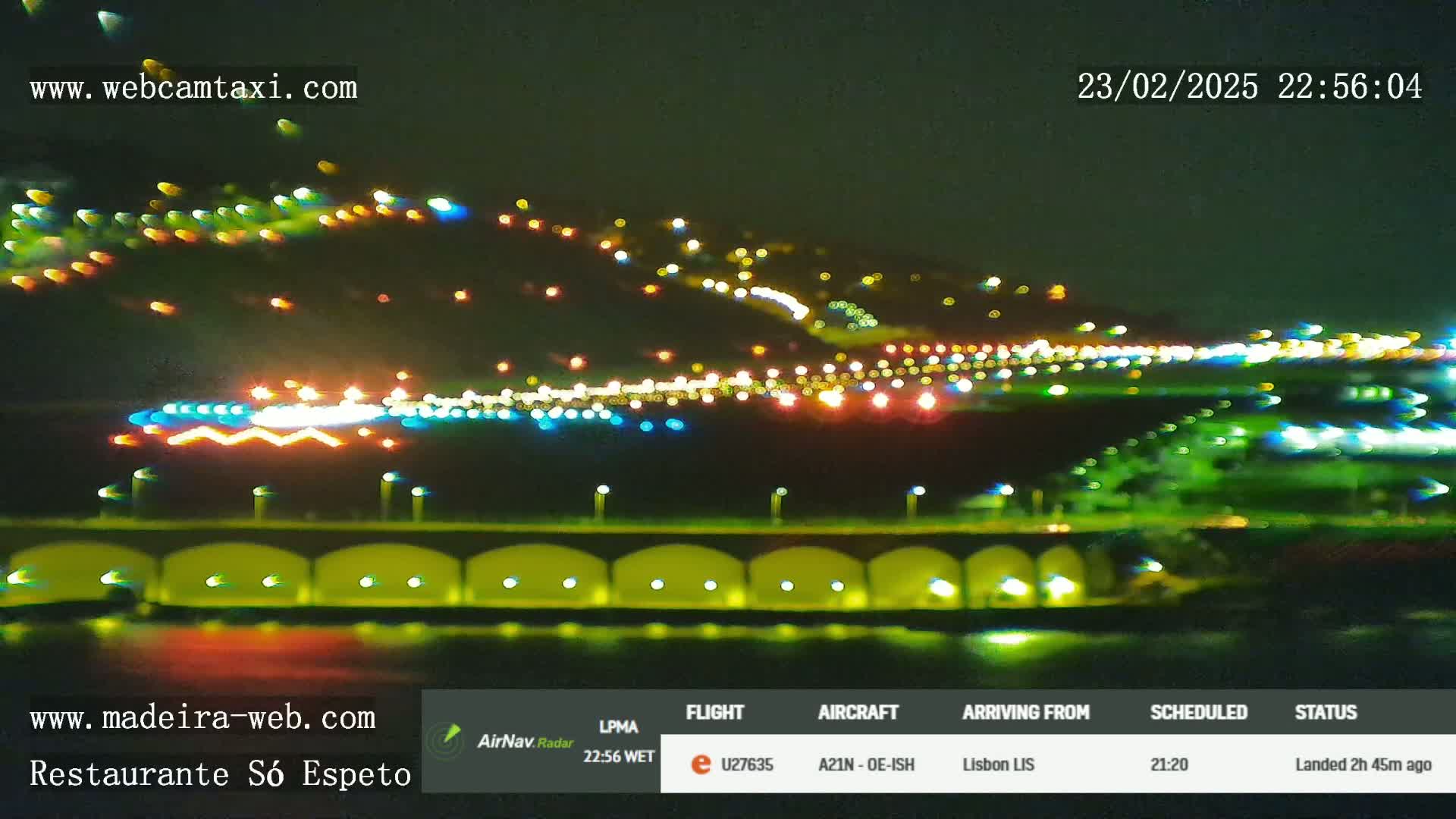 A nighttime, blurry long shot of a brightly lit airport runway and surrounding area, with city lights visible in the distance.