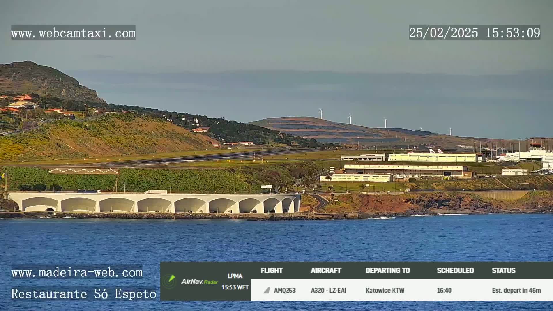 An airport runway and surrounding buildings are shown on a clear day next to a body of water with a coastline.