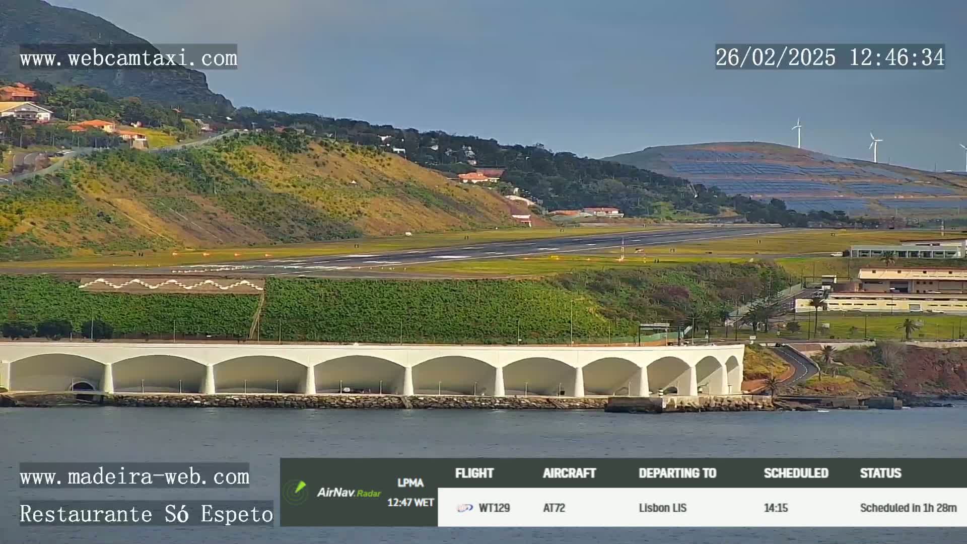 An airport runway is visible from a waterfront vantage point on a sunny day, with a curved structure featuring multiple arches in the foreground.
