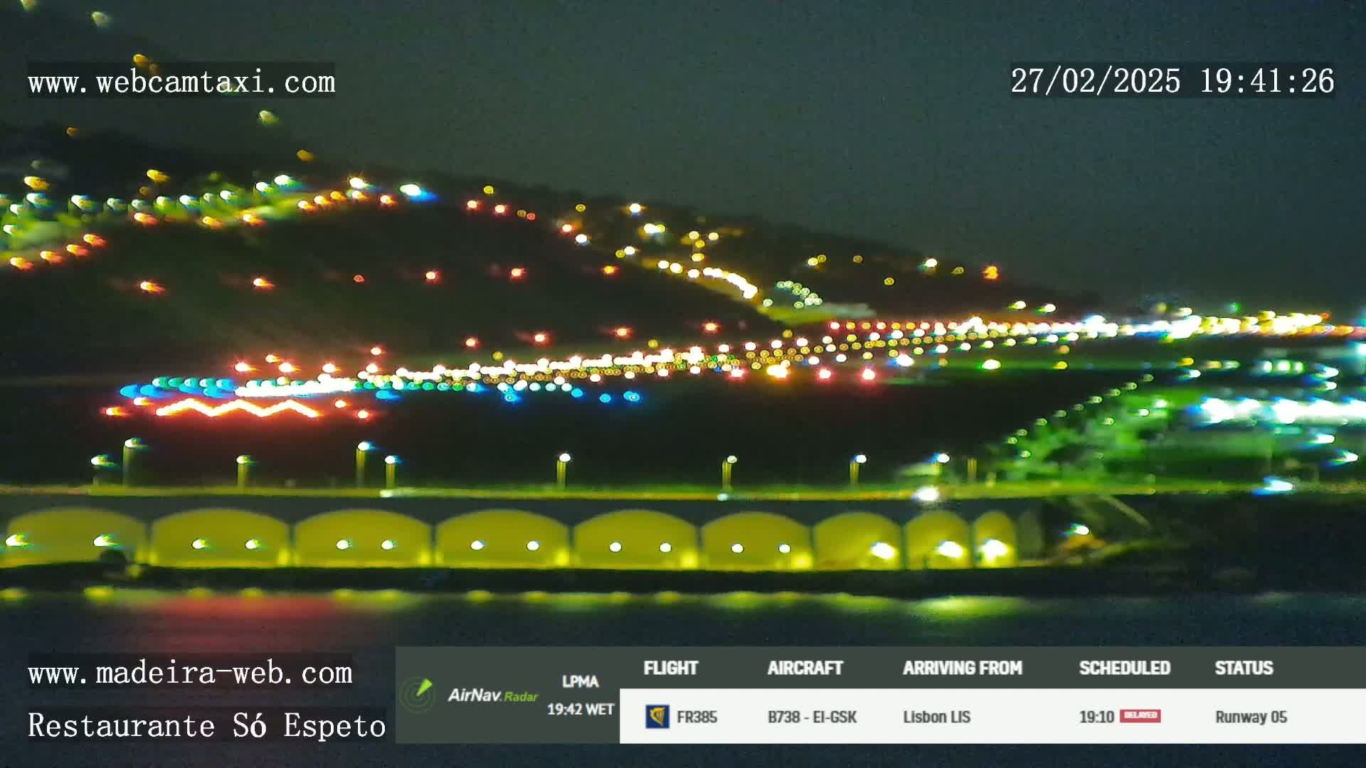 A nighttime view of a brightly lit airport runway and surrounding area with city lights on a hill in the background.