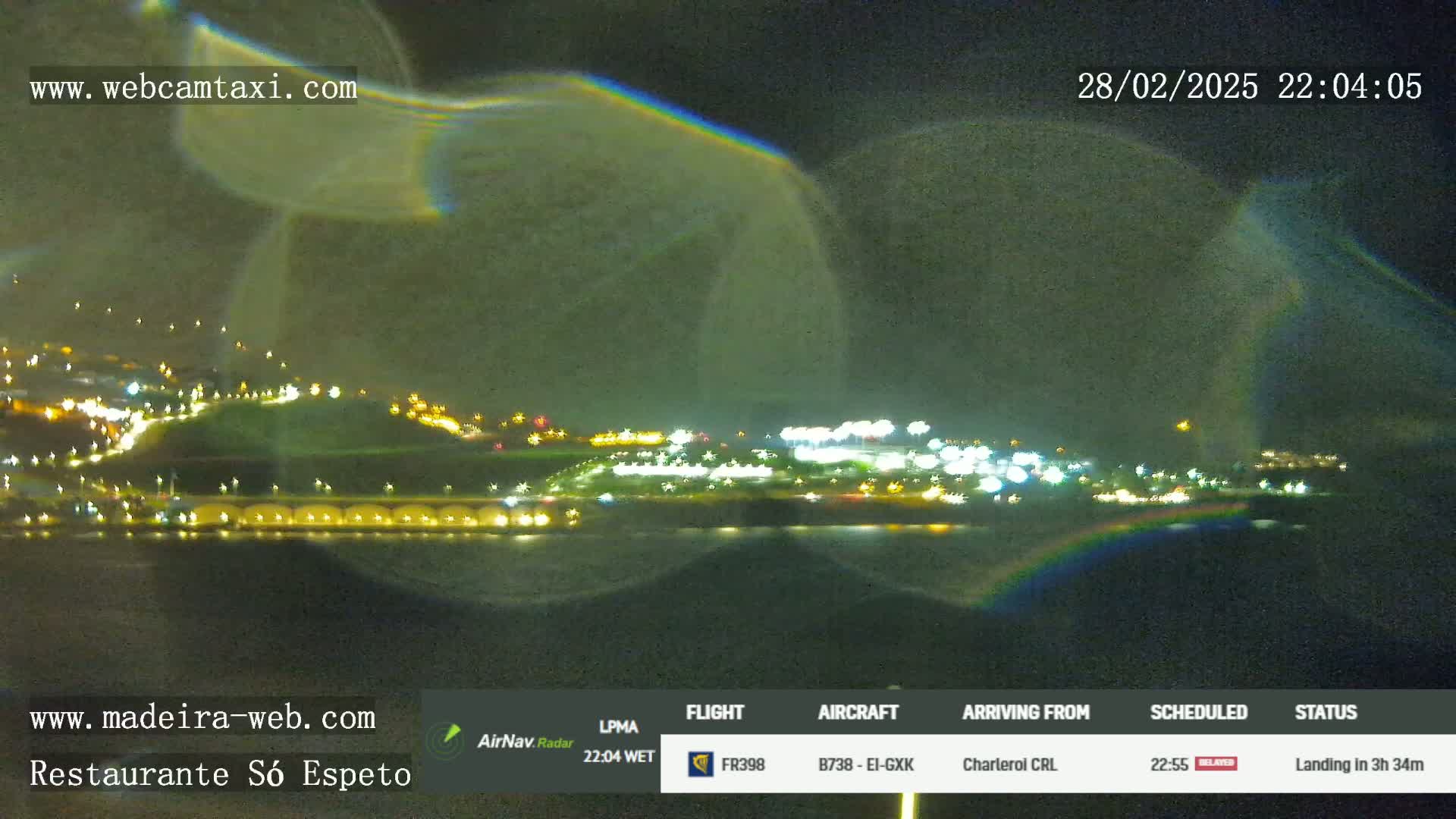 A nighttime view of an airport runway and surrounding area illuminated by lights, with a faint rainbow-like effect in the sky.