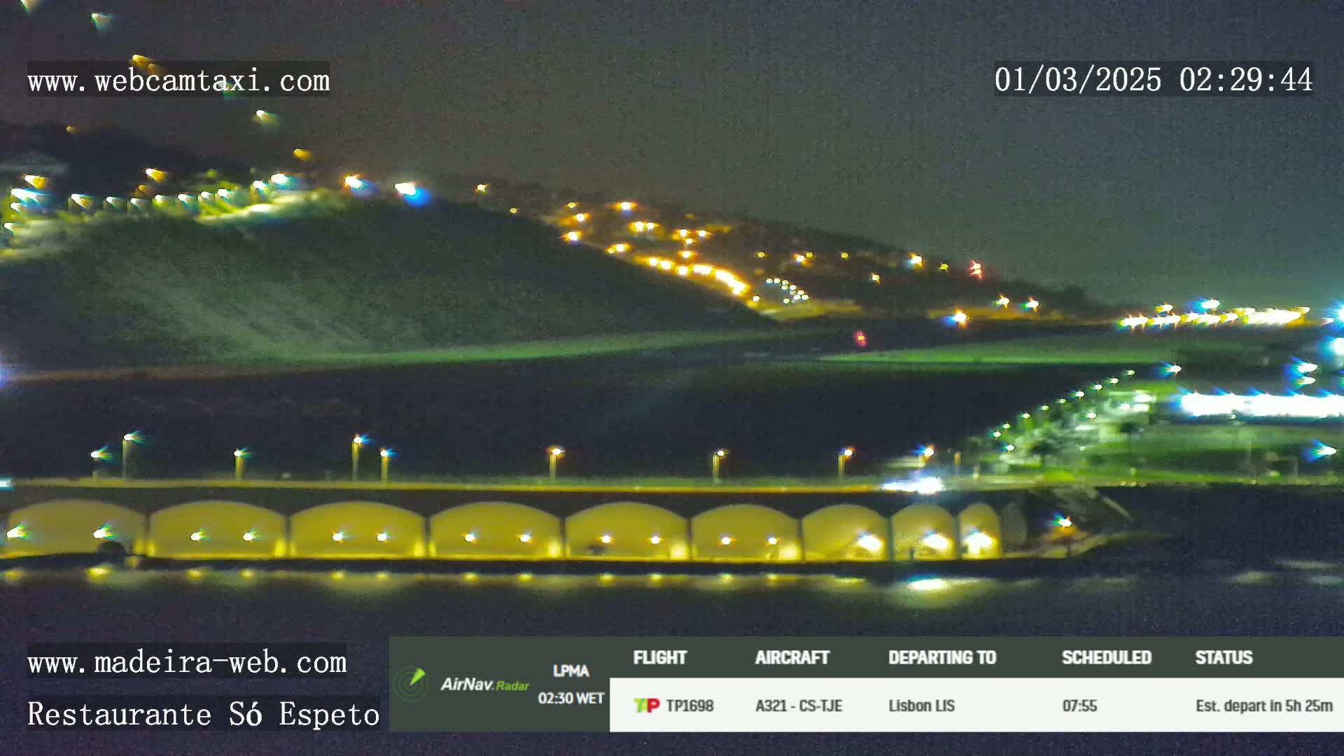 A nighttime view of a coastal town with hills lit by many lights, seen across a body of water next to a bridge with lights under it.