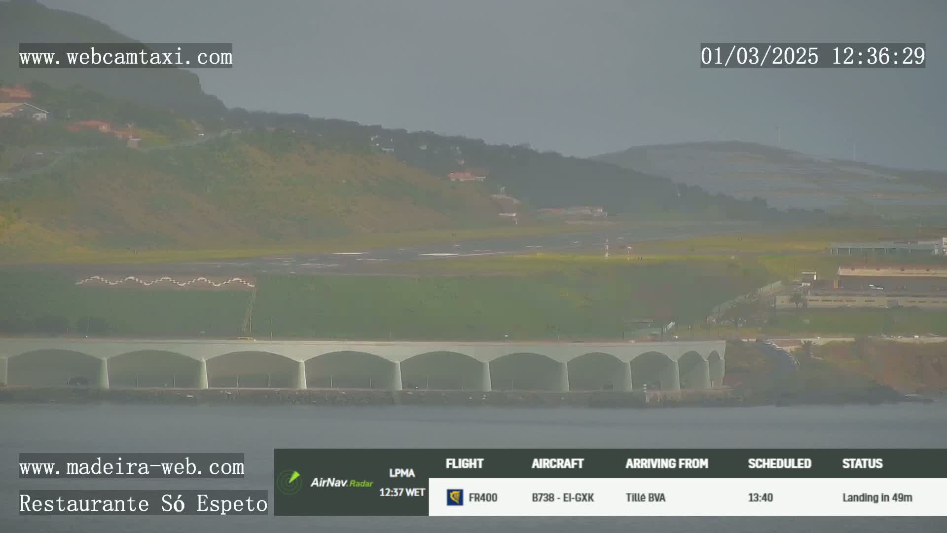 A hazy view shows an airport runway and a long, arched structure by the water, with hills in the background.