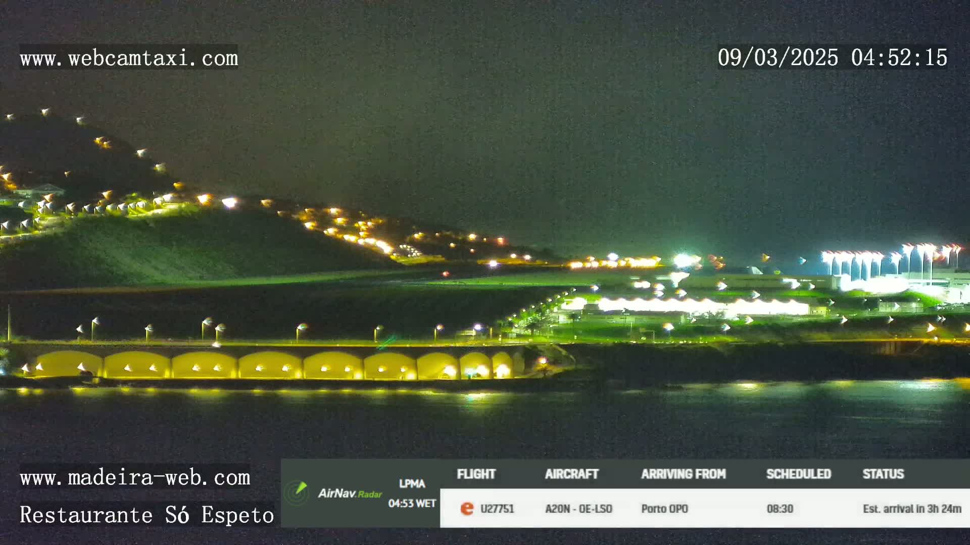 A nighttime view of a coastal airport with illuminated buildings and a hill in the background, under clear skies.