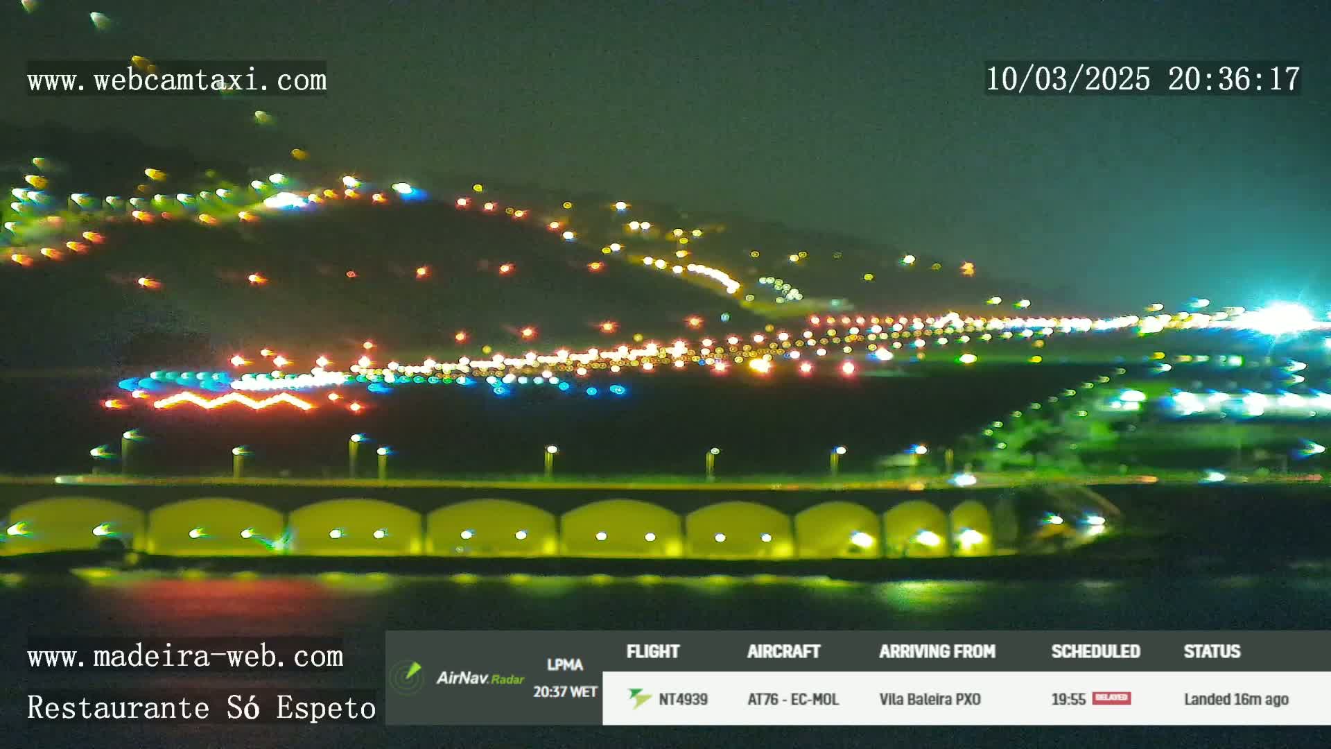 A nighttime, blurry long shot of an airport runway and surrounding lights, with a hill in the background.