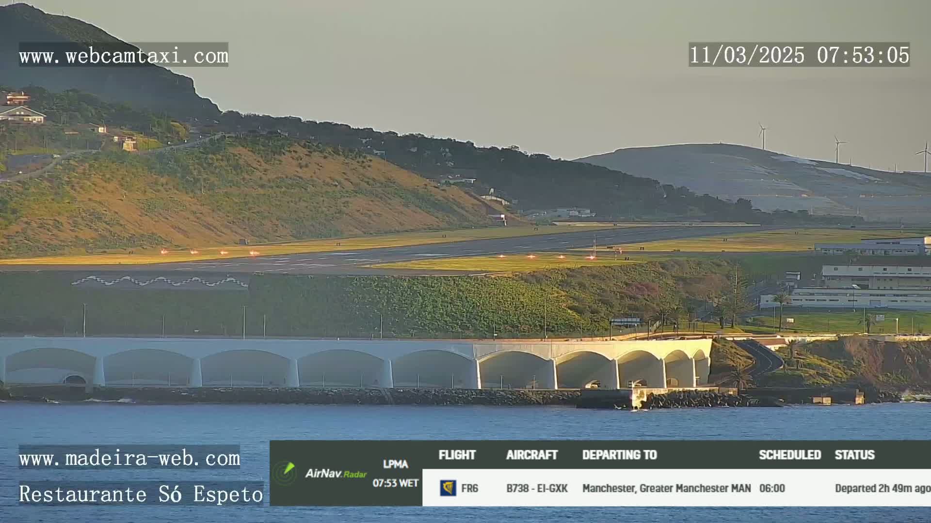 An airport runway with a plane taking off is shown in the early morning light, with a coastal area and a body of water visible in the foreground.