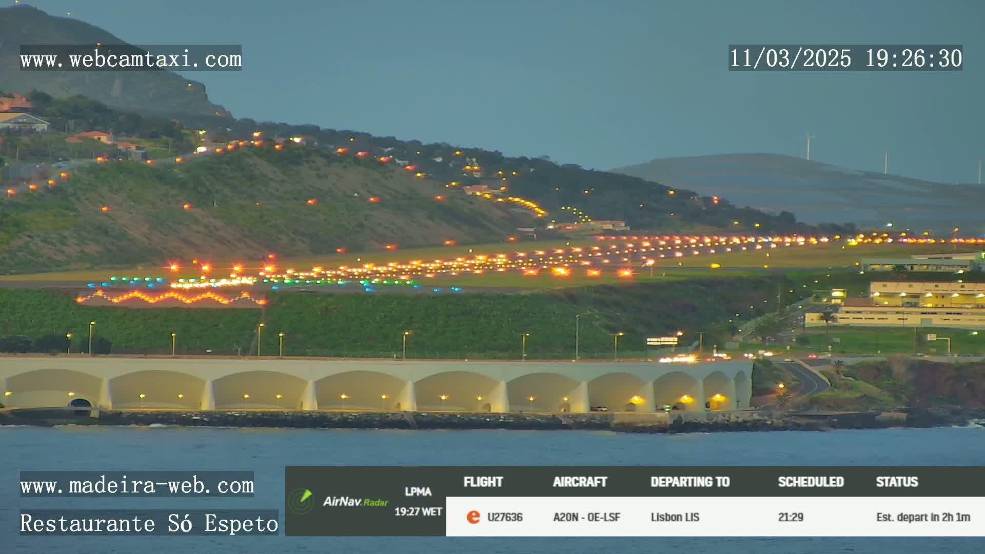 A brightly lit airport runway at dusk, situated along a dark hillside next to a calm body of water.