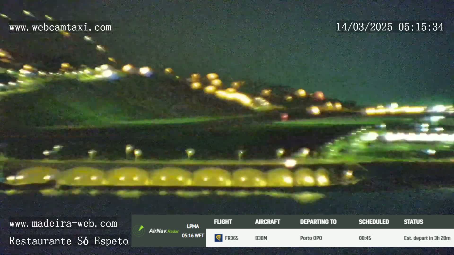 A nighttime view shows a blurry image of lights on a hillside and a row of illuminated structures near a body of water.