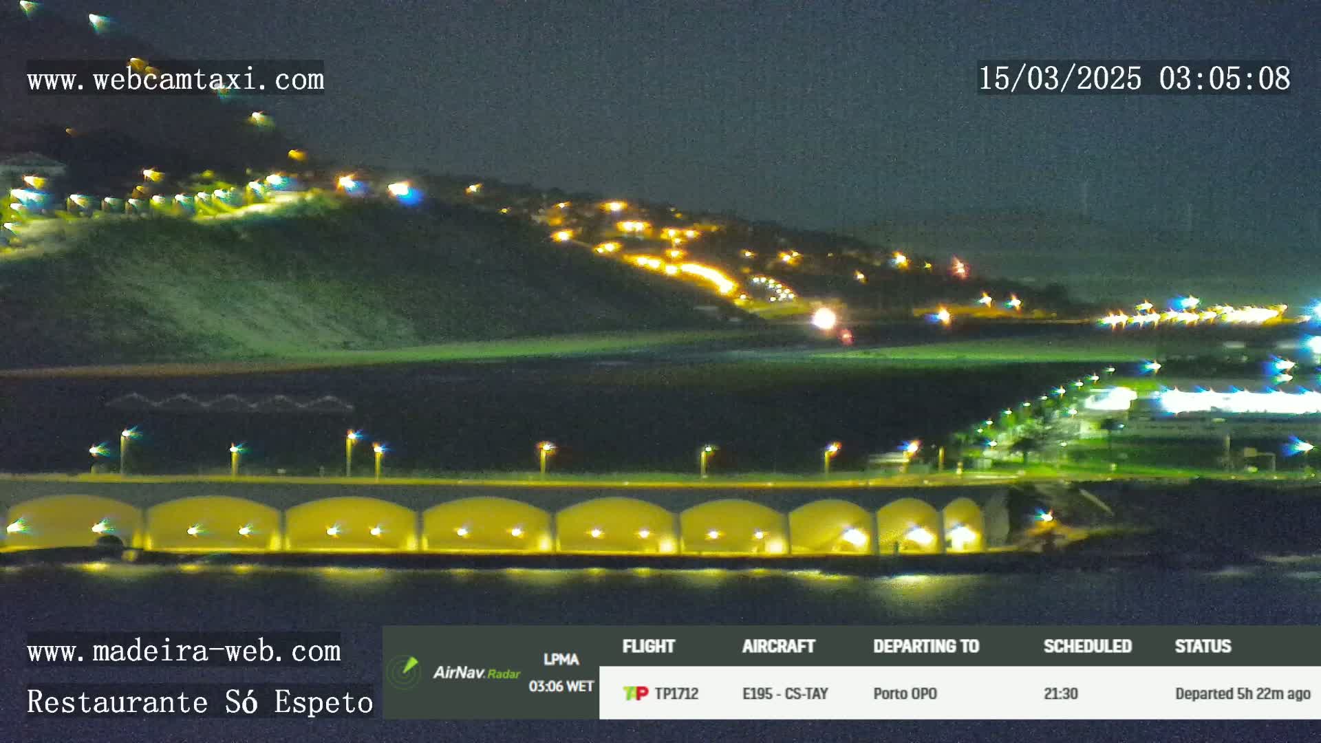 A nighttime coastal scene shows a hilltop with illuminated buildings overlooking a beach and a lit arched bridge extending into the water.