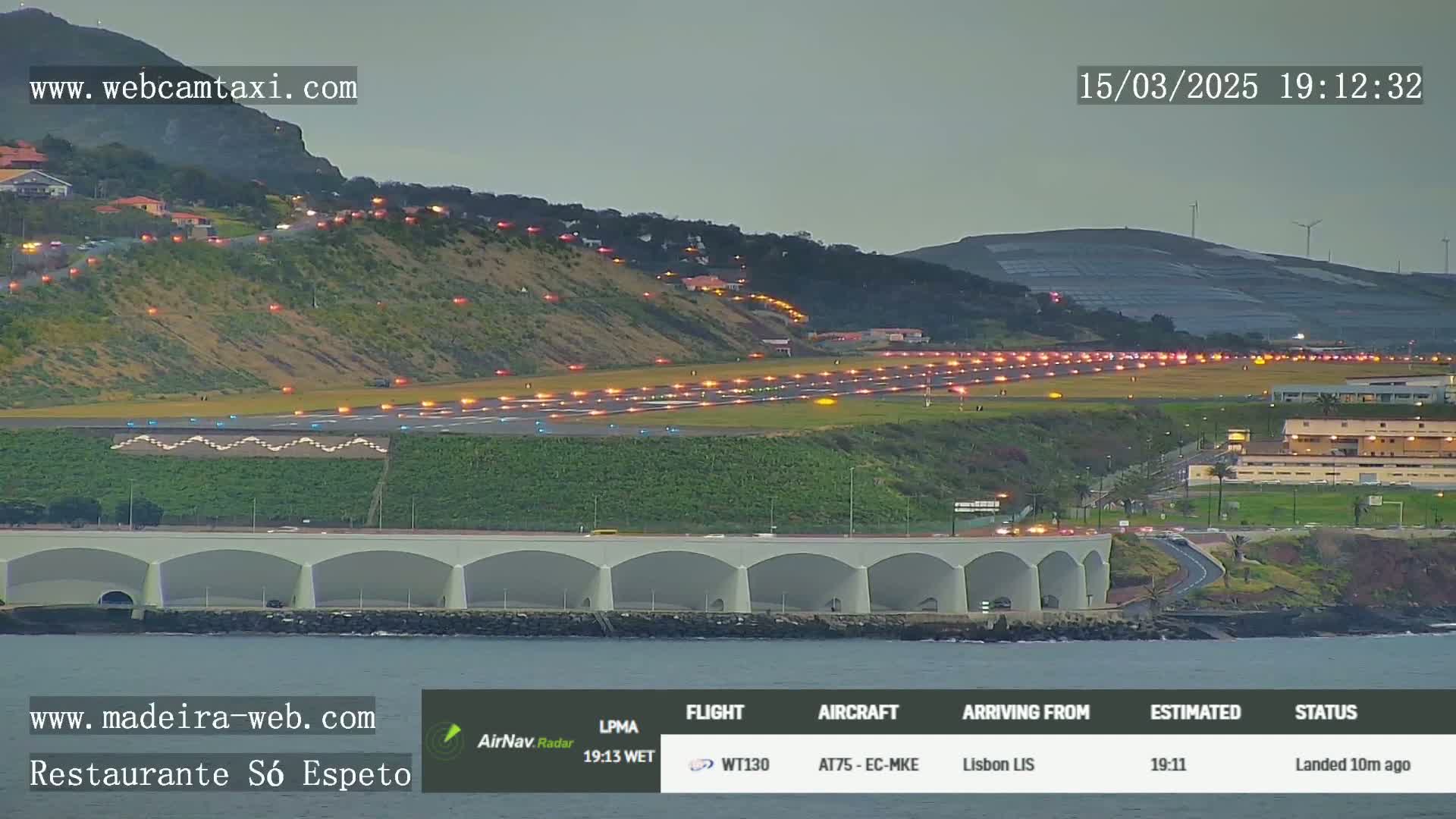 An airport runway at dusk, illuminated by runway lights, is adjacent to a coastal road and a bridge-like structure over the water.