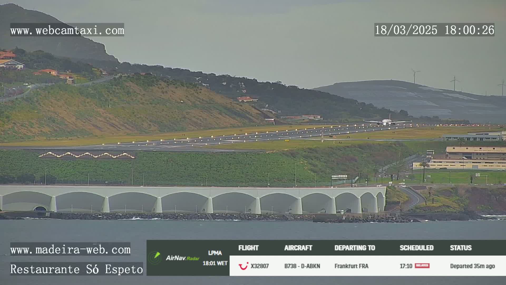 An airplane is taking off from an airport runway beside a body of water, under overcast skies.
