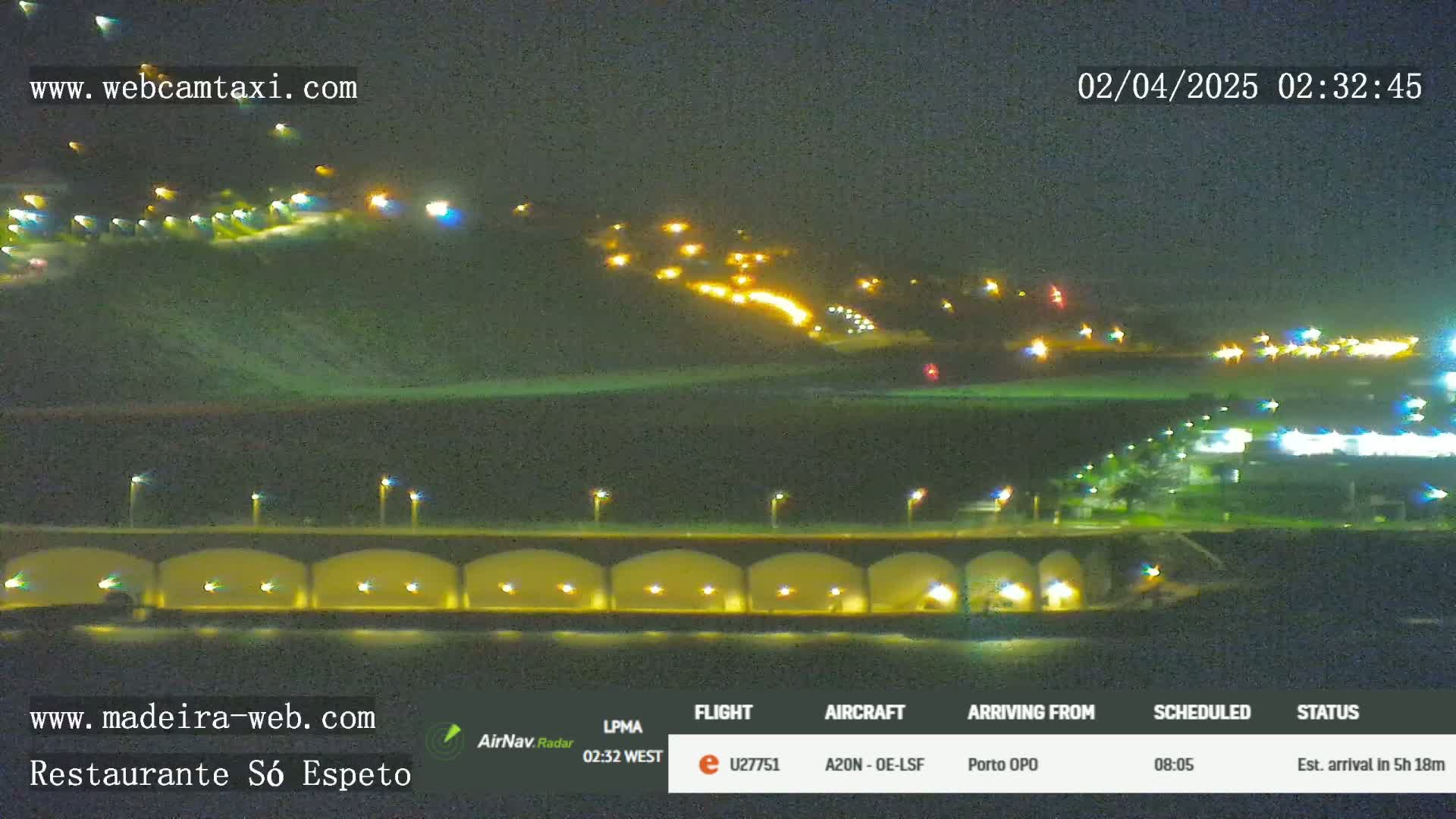 A nighttime view of a coastline with illuminated buildings on a hill and a bridge with arched underpasses, under a clear sky.