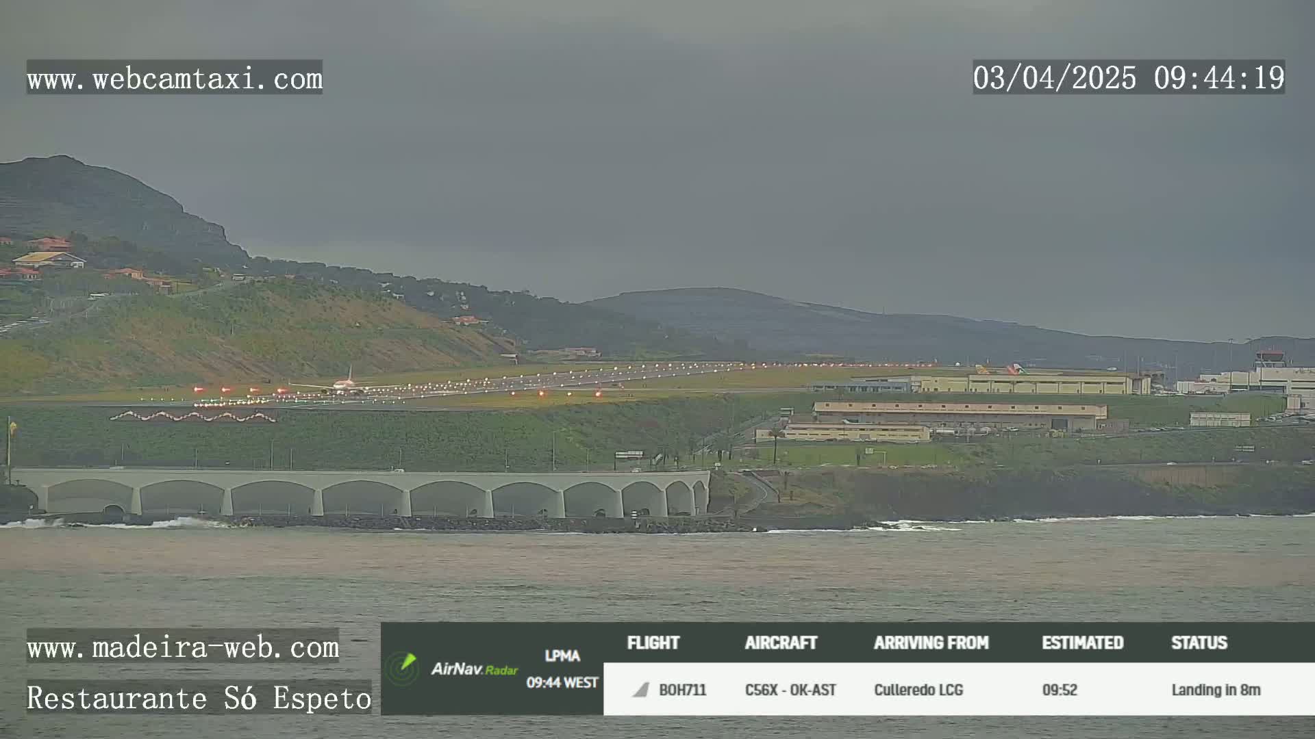 An airplane is landing on a runway near an ocean under an overcast sky.